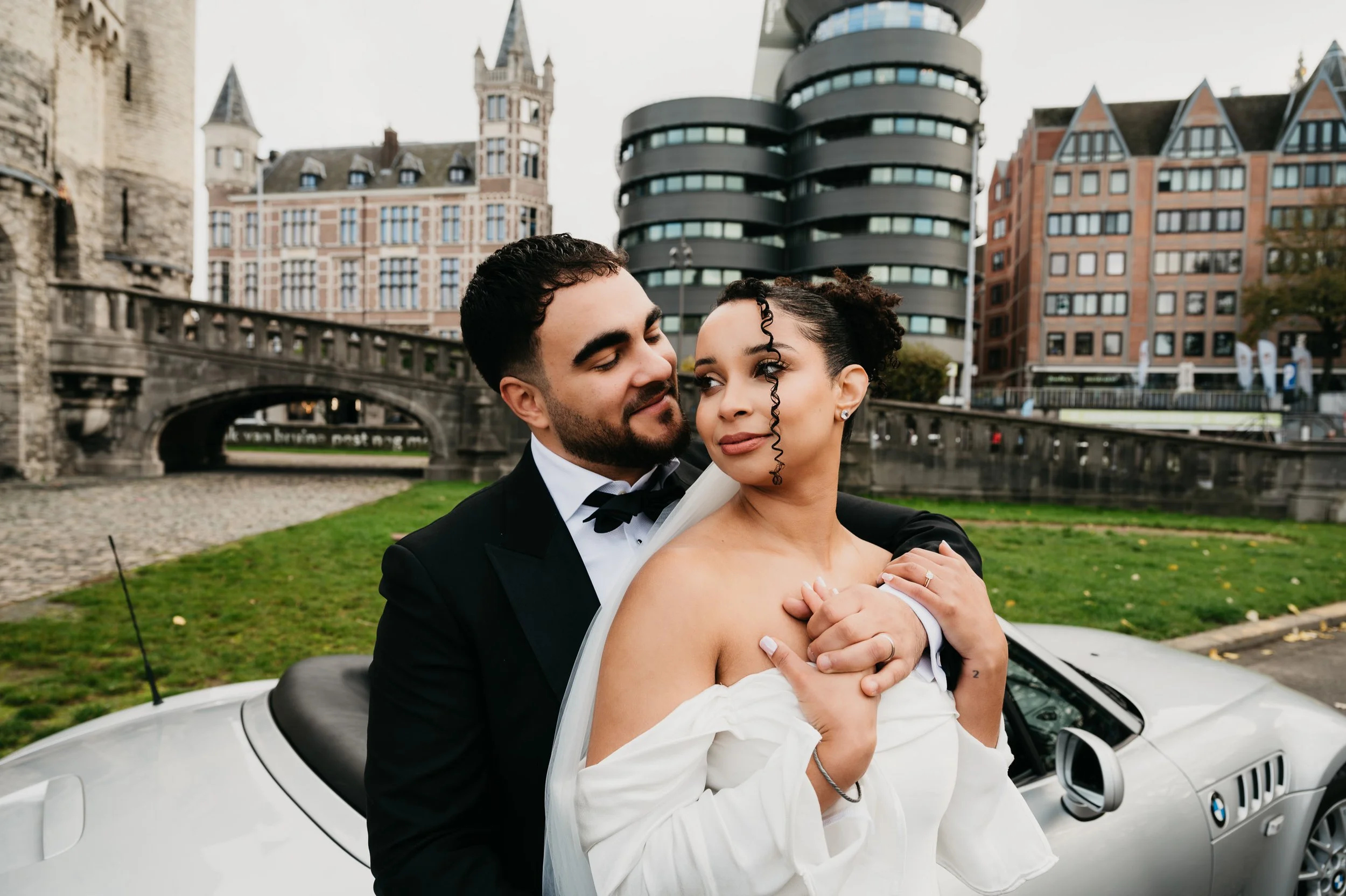 A newlywed couple in wedding attire embracing in front of a silver convertible car with a cityscape background of modern and historic buildings.