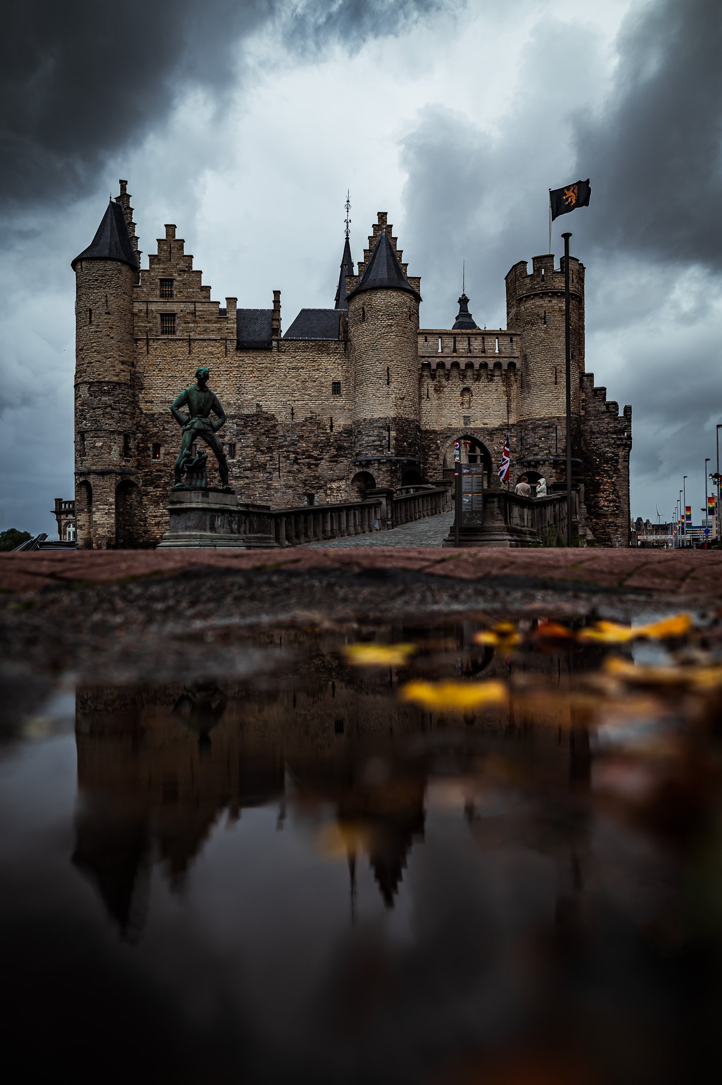A historic castle with turrets and towers under a cloudy sky, with a statue of a man in front and a reflection on a wet surface in the foreground.