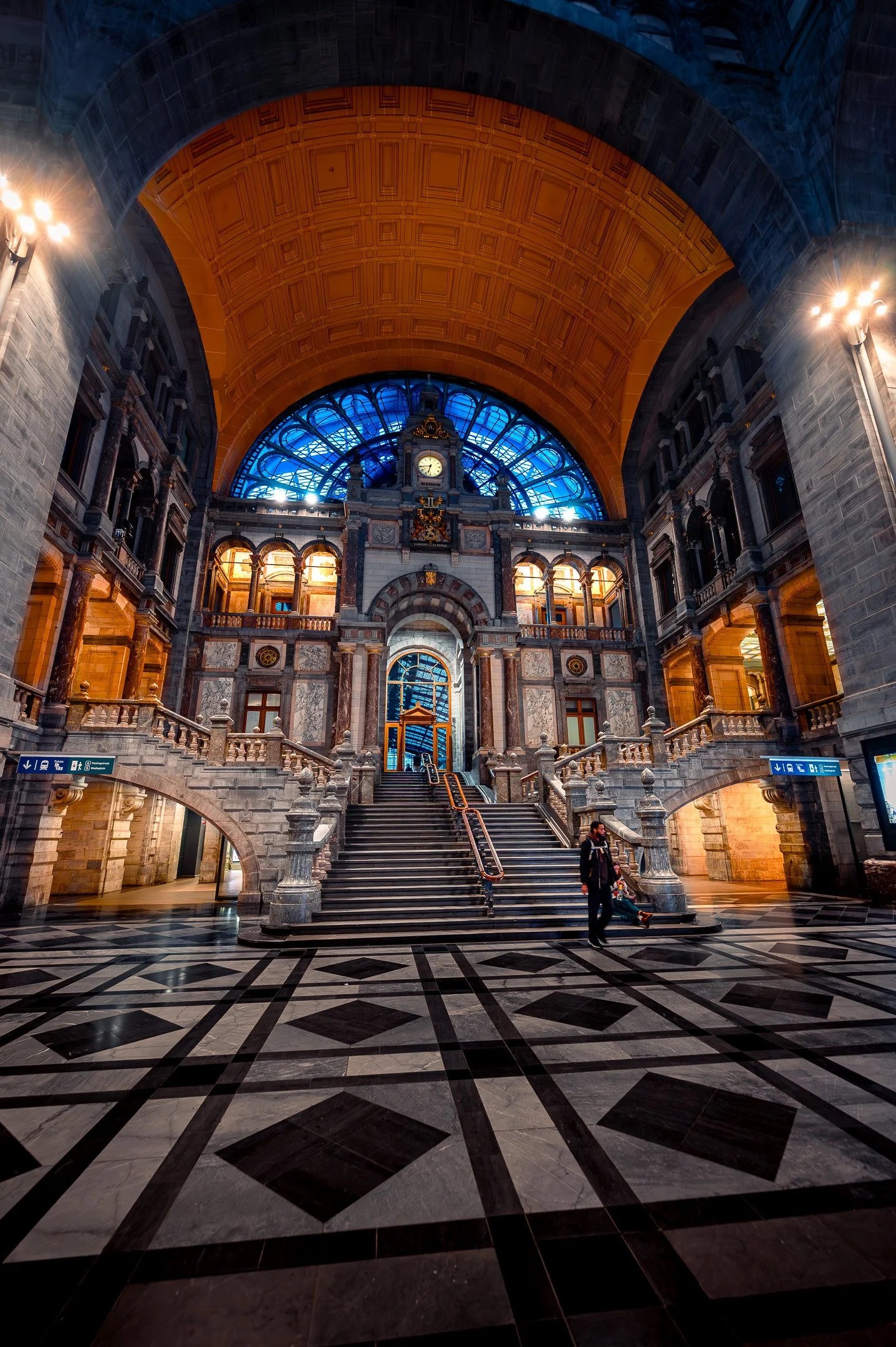 Grand interior of a historic train station with a grand staircase, marble floors, arches, ornate architecture, large clock, and blue glass ceiling.