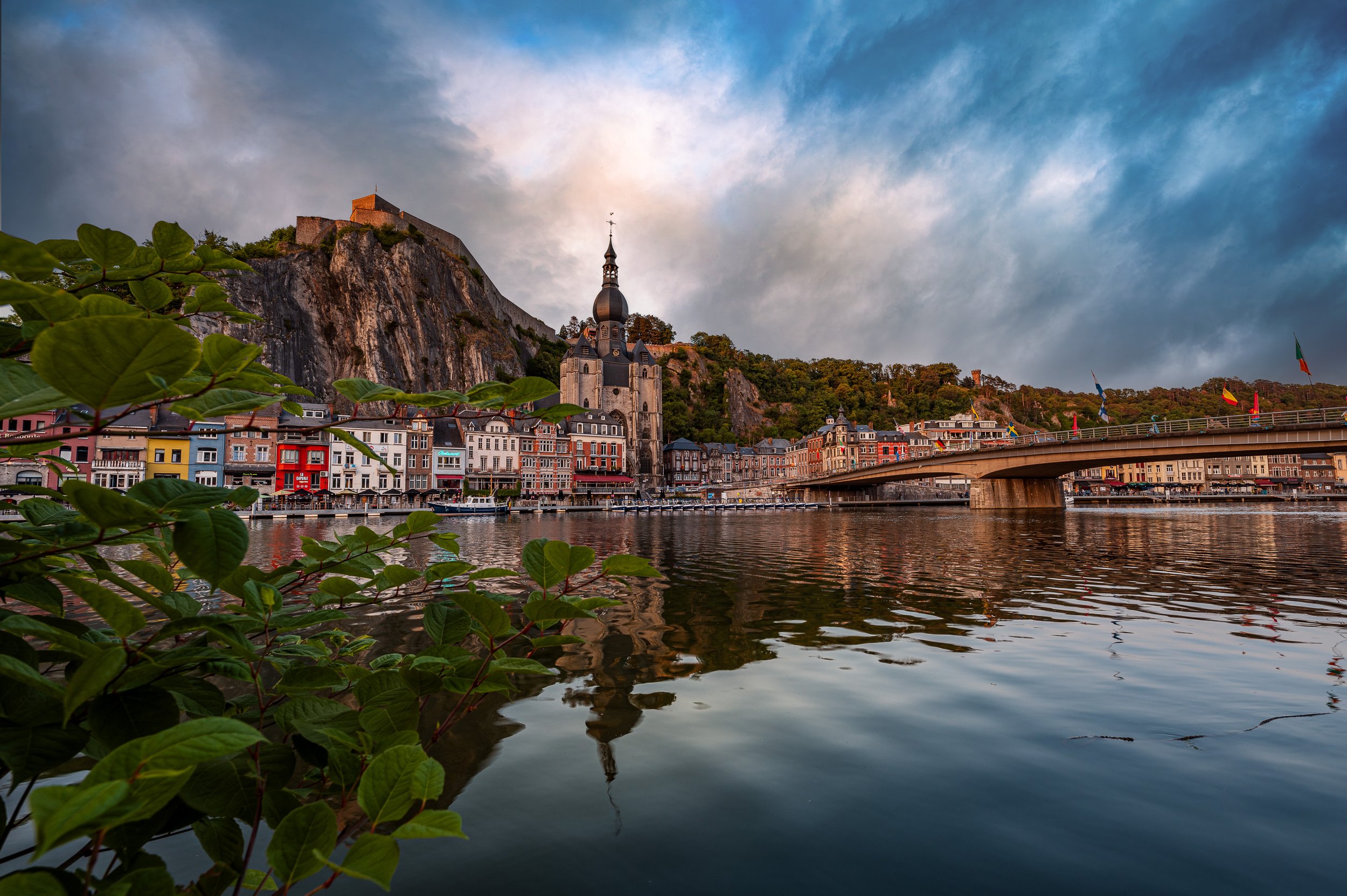A scenic view of a historic European city with colorful buildings along a river, a church with a tall spire, a stone bridge, and a fortress on a hill under a partly cloudy sky.