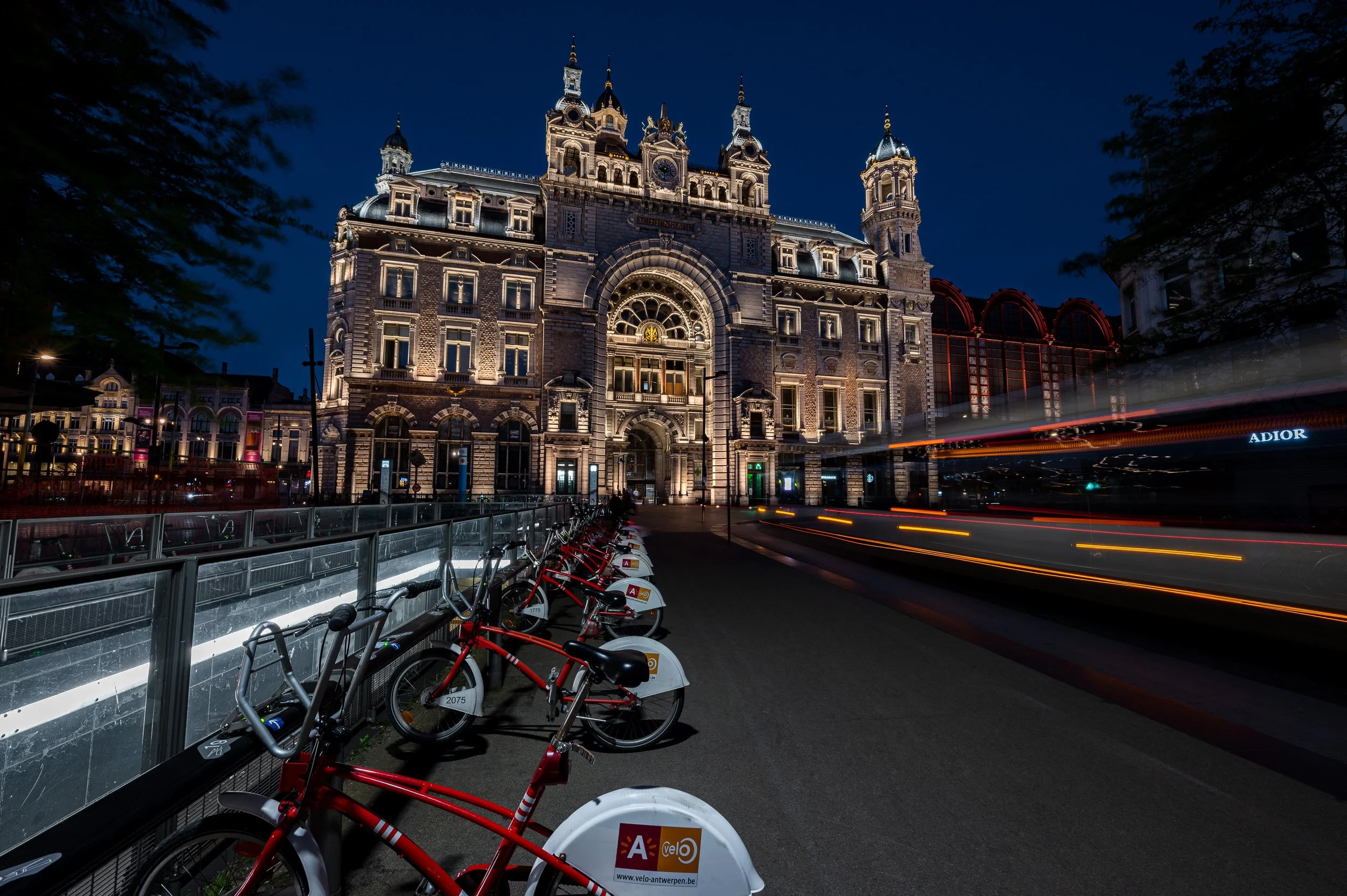 Nighttime view of a historic building with illuminated ornamentation, fronted by a row of red and black bicycles parked along the sidewalk and streaks of moving vehicle lights on the street.