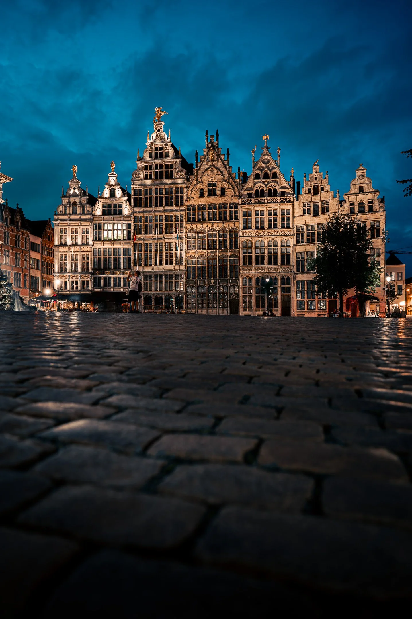 Nighttime view of historical buildings with ornate facades on a cobblestone square, illuminated by streetlights, with a dark, cloudy sky overhead.