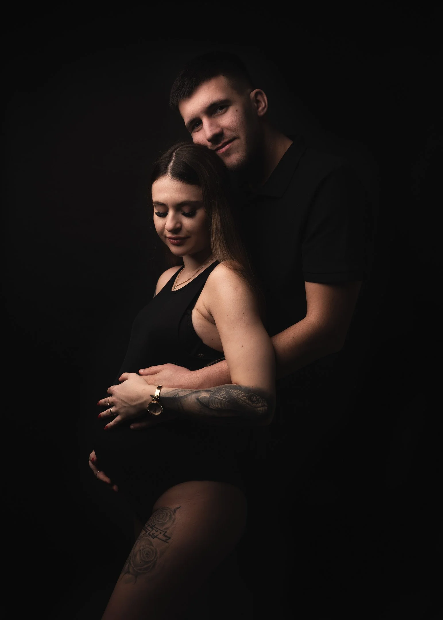 A pregnant woman with tattoos on her arm and thigh, wearing a black sleeveless top, stands with her eyes closed while a man stands behind her, gently holding her belly with his arms around her, against a black background.