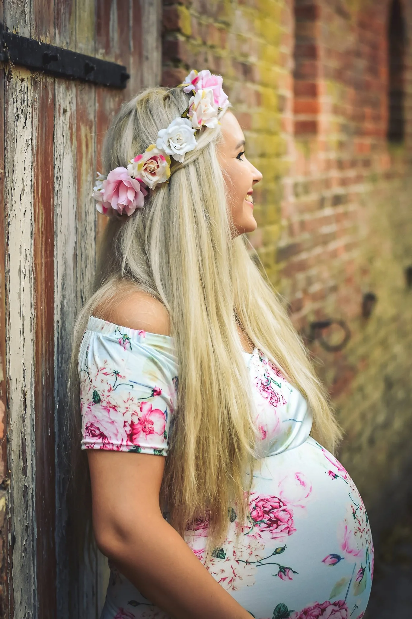 Pregnant woman with long blonde hair wearing a floral dress and a flower crown, leaning against a weathered brick wall, smiling.