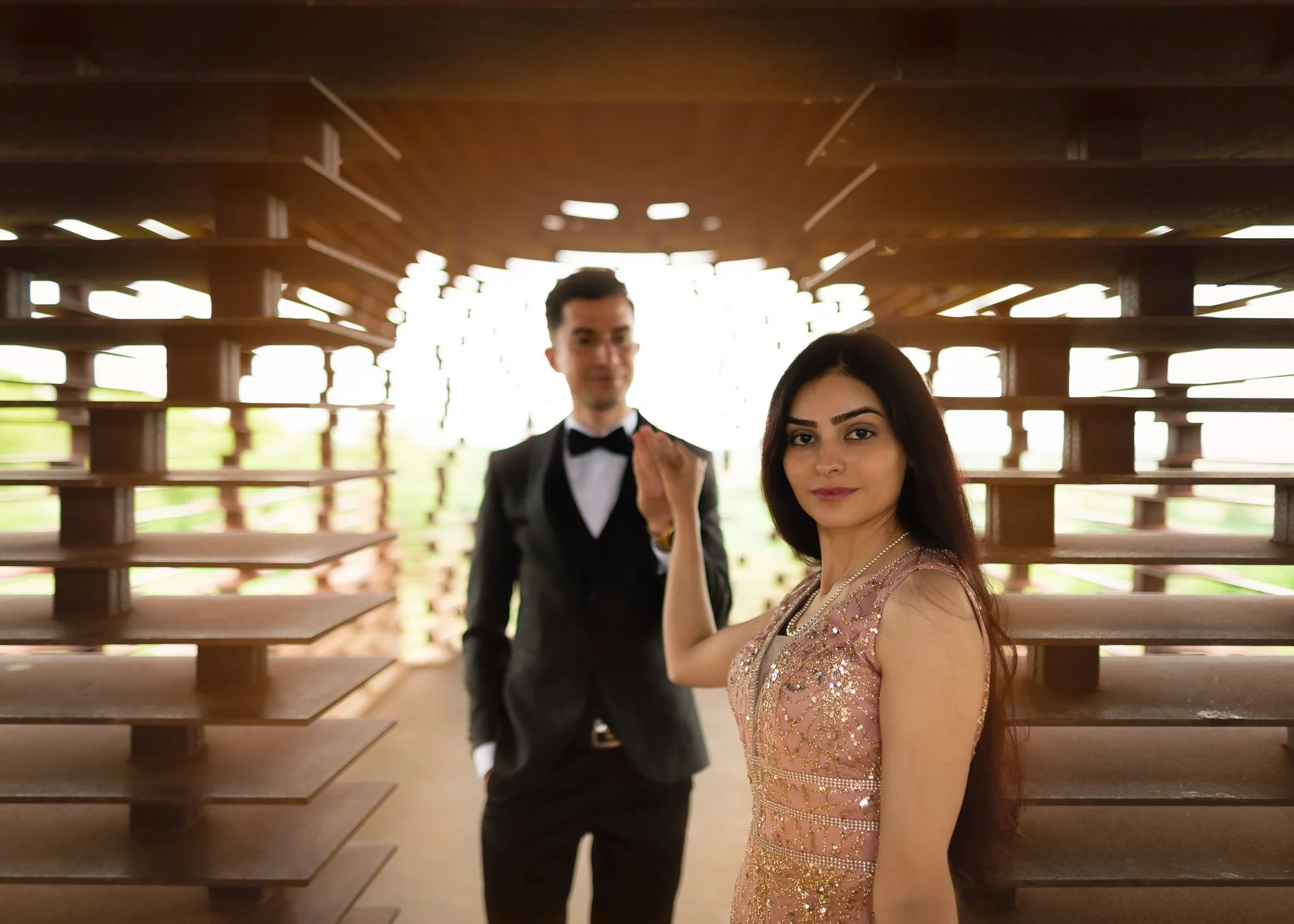 A woman in a pink, beaded dress in the foreground, and a man in a black tuxedo with a bow tie in the background, standing inside a modern wooden structure with horizontal slats, with bright natural light behind them.