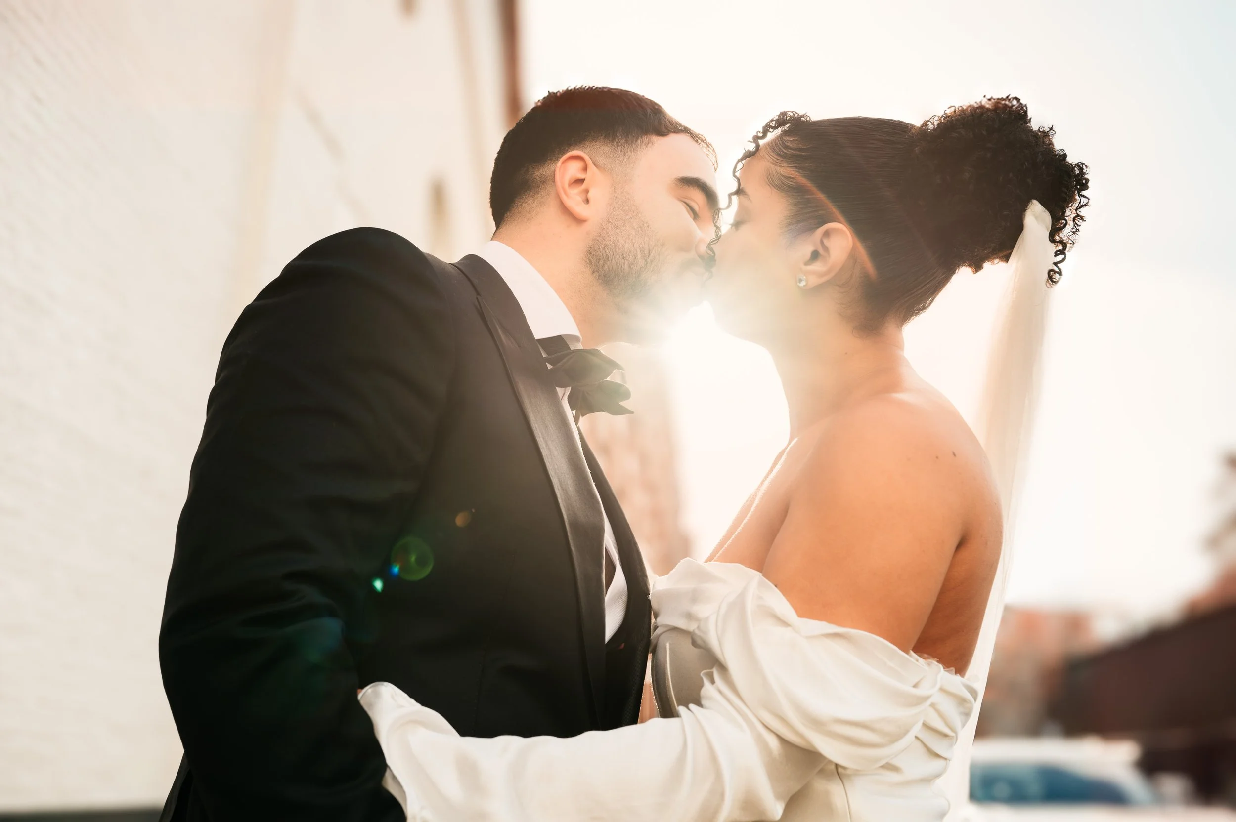 A newlywed couple in wedding attire sharing a kiss outdoors with the sun shining behind them.