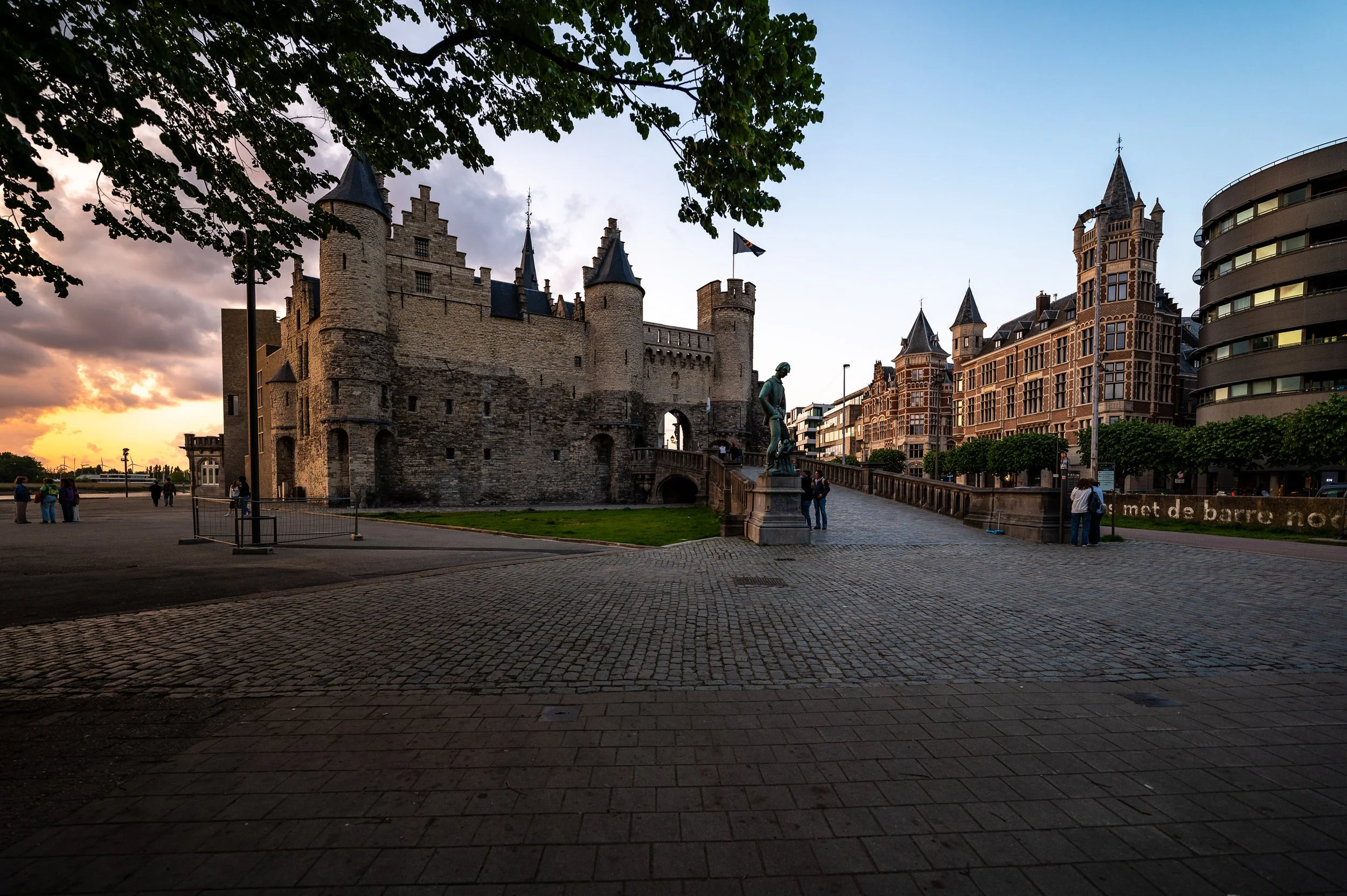 A stone castle on a cobblestone street with a statue in front, modern buildings on the right, and a sunset behind the castle.