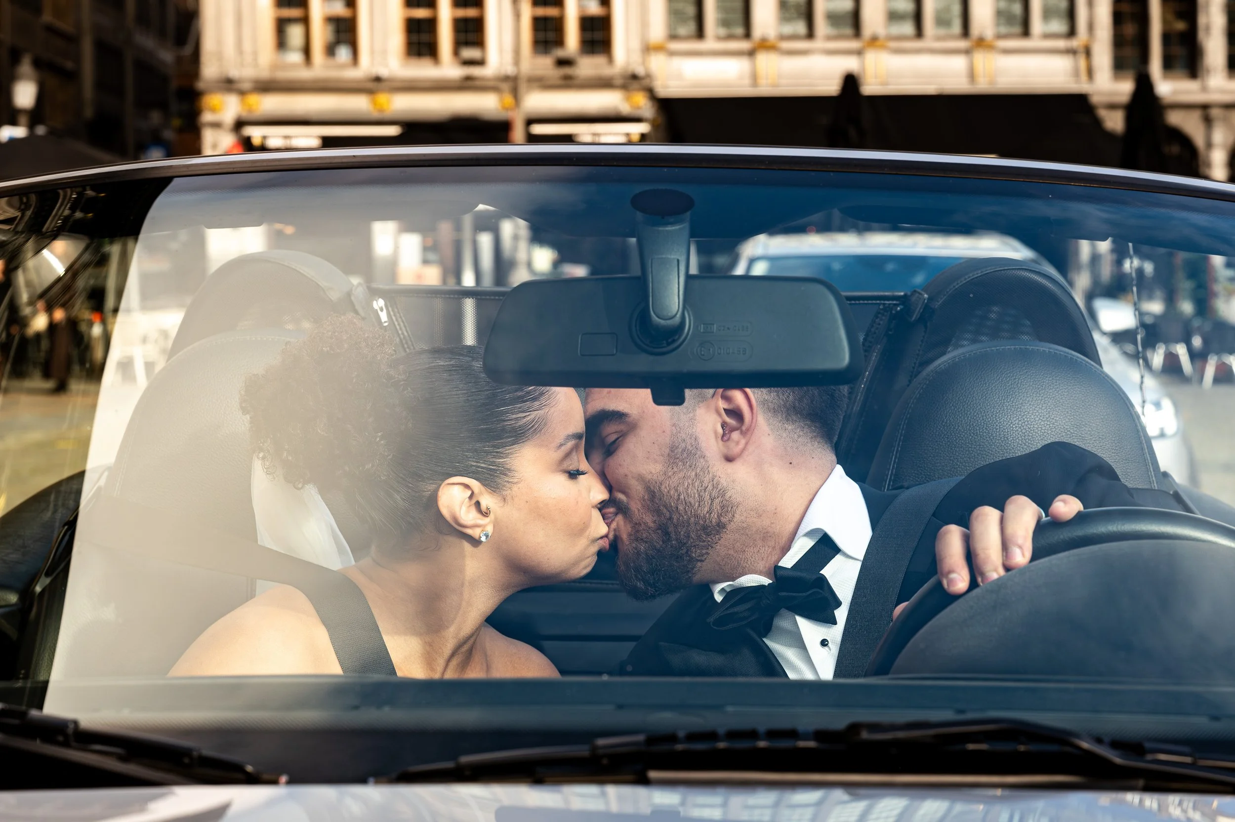 A couple dressed in wedding attire sharing a kiss inside a car.