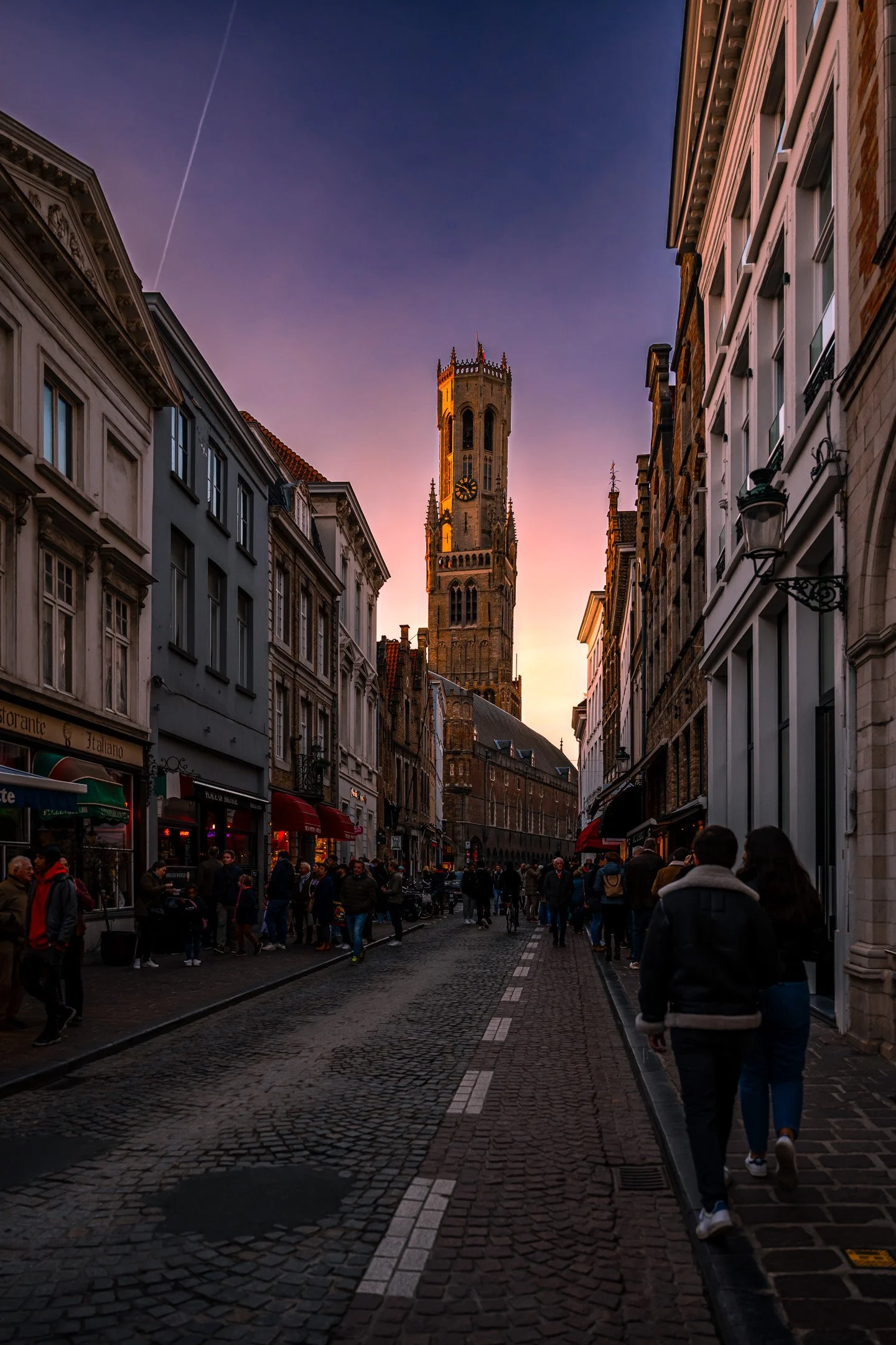 A city street scene at sunset with a tall clock tower in the background, people walking on cobblestone pavement, and buildings with shops and cafes lining both sides.