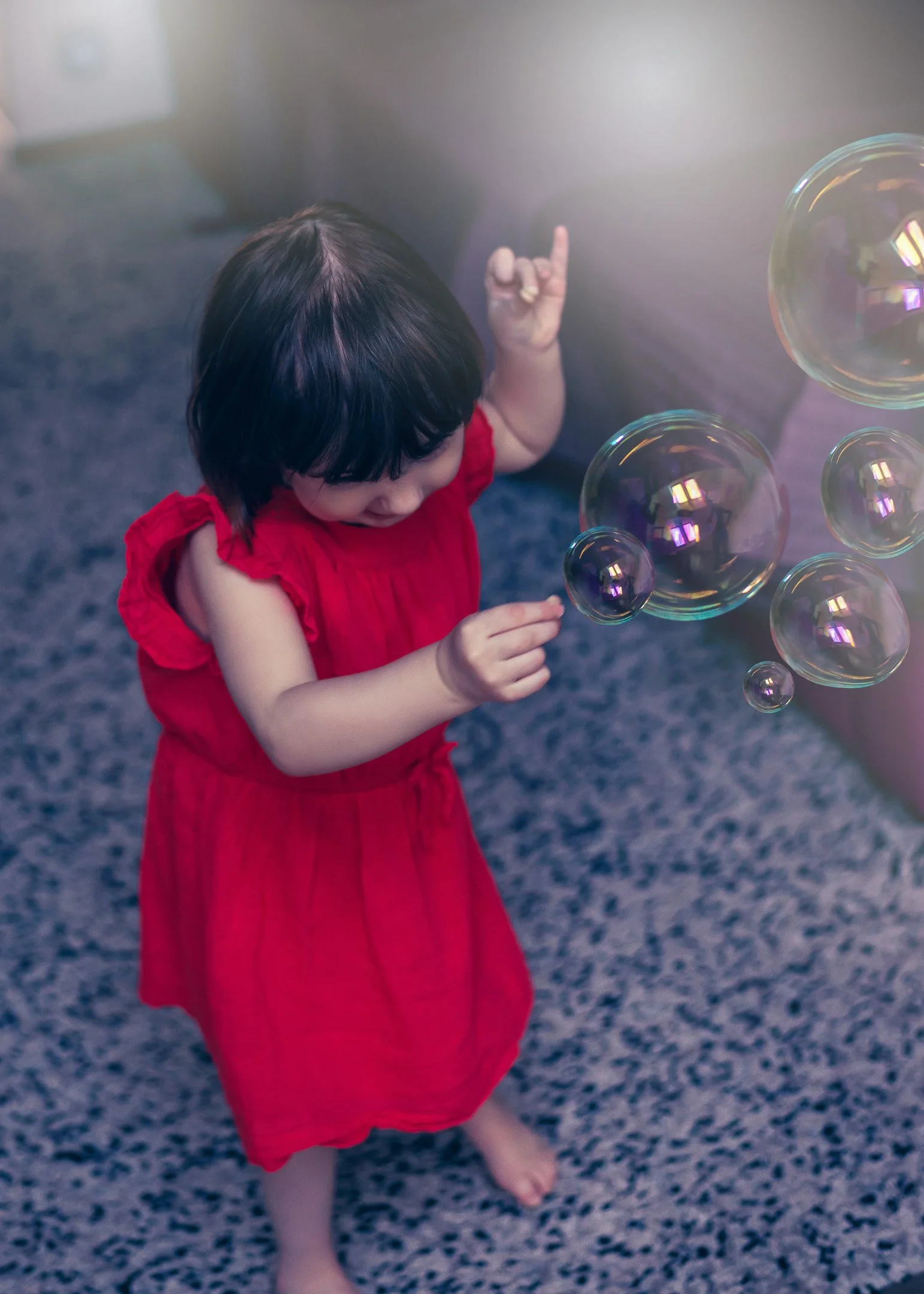 A young girl in a red dress playing with soap bubbles indoors on a gray carpeted floor.