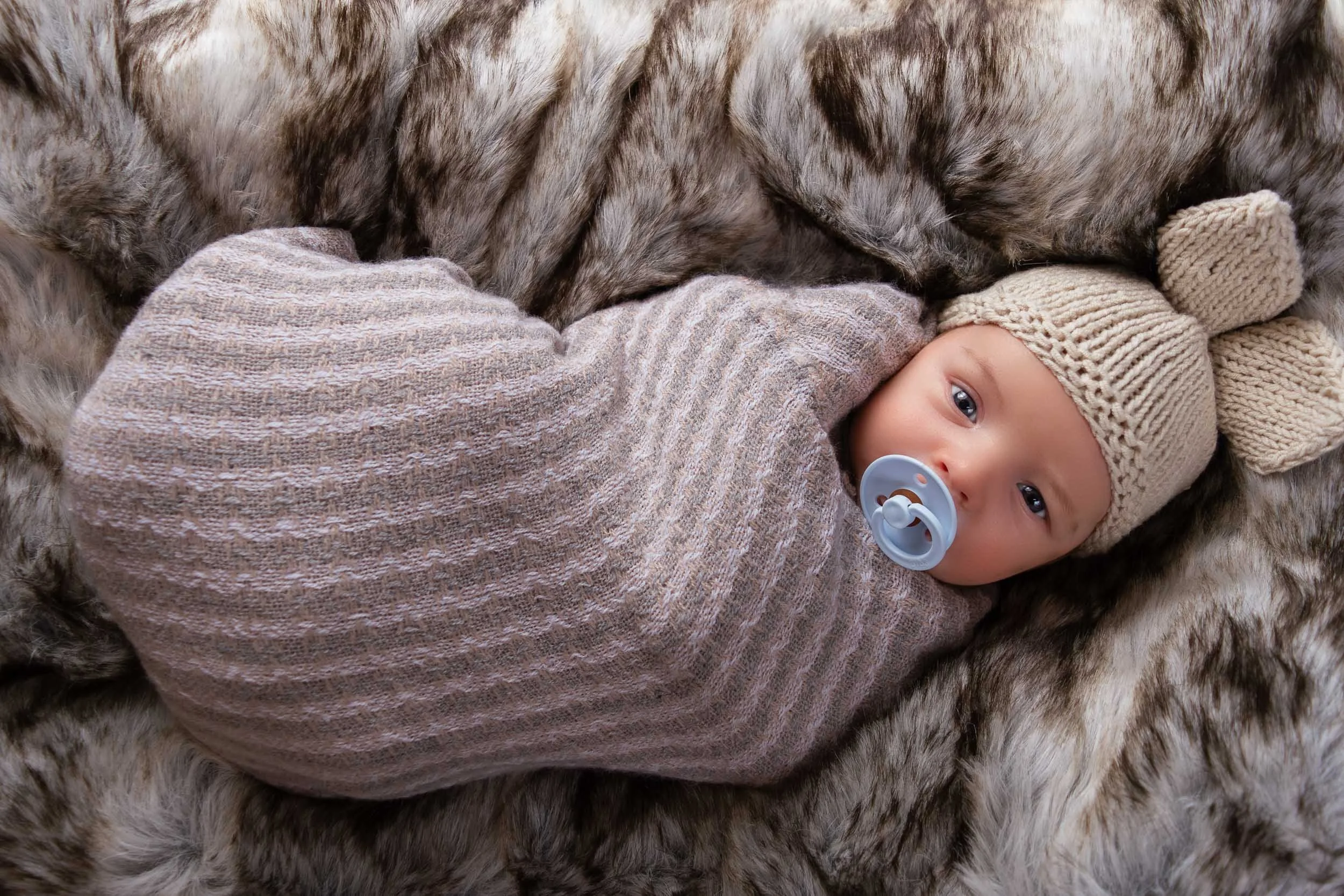 A baby lying on a fur blanket wearing a beige knit hat with bear ears and a pacifier, wrapped in a cozy, striped blanket.