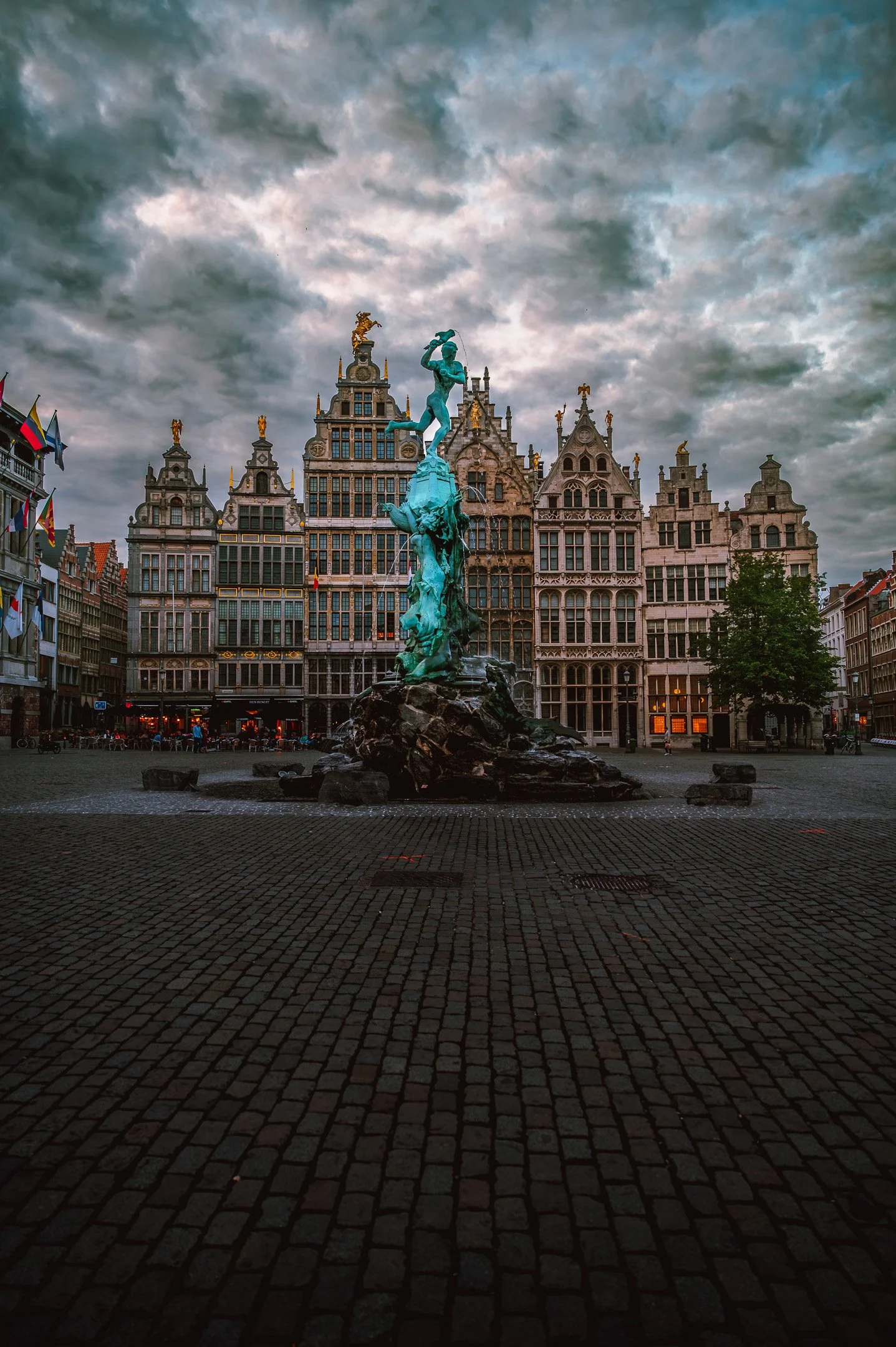 A historic square featuring a statue with a stylized European cityscape backdrop and a moody cloudy sky.