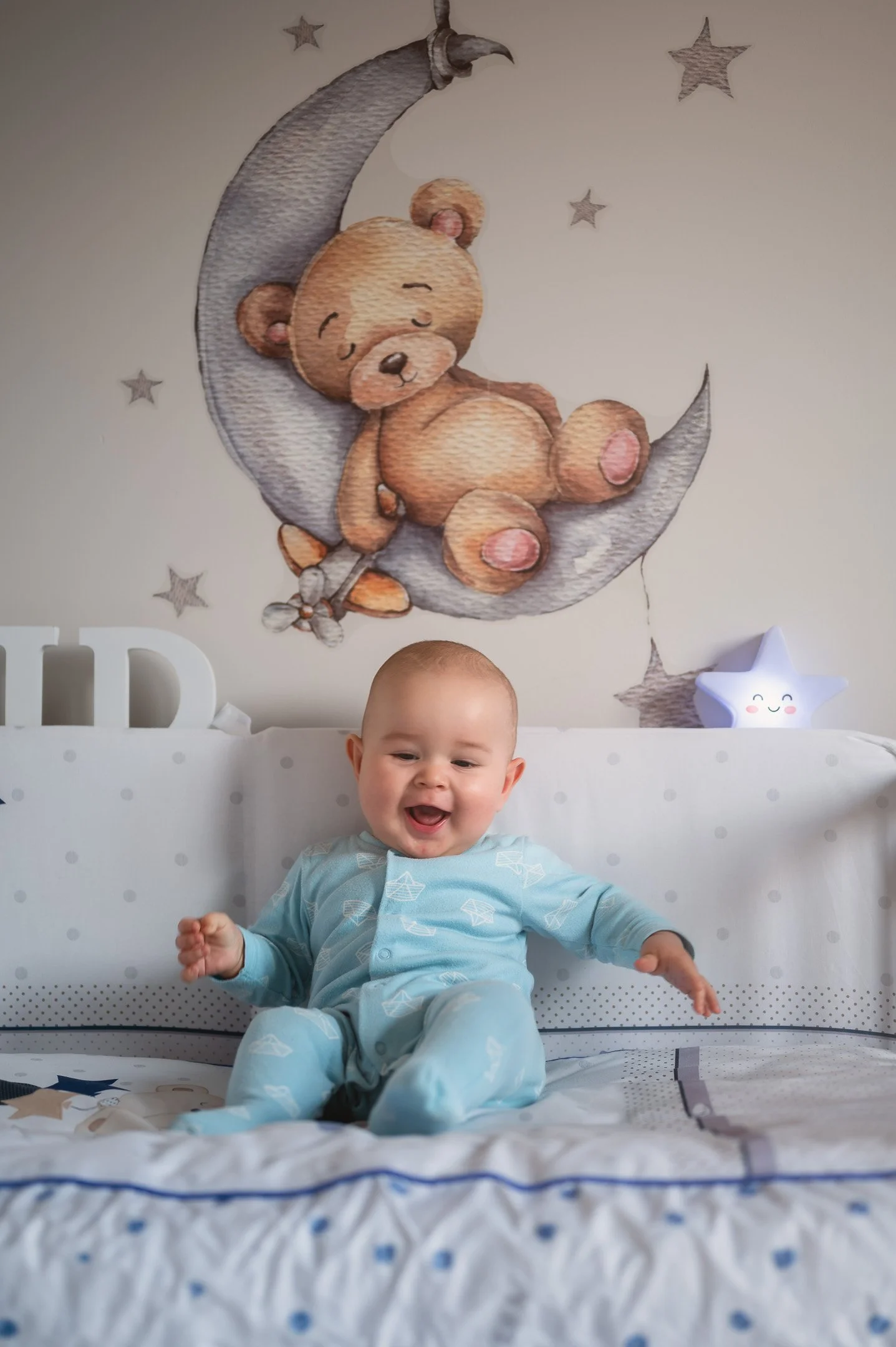 A smiling baby in a light blue pajama set sitting on a bed with a teddy bear and star-themed decor in the background.