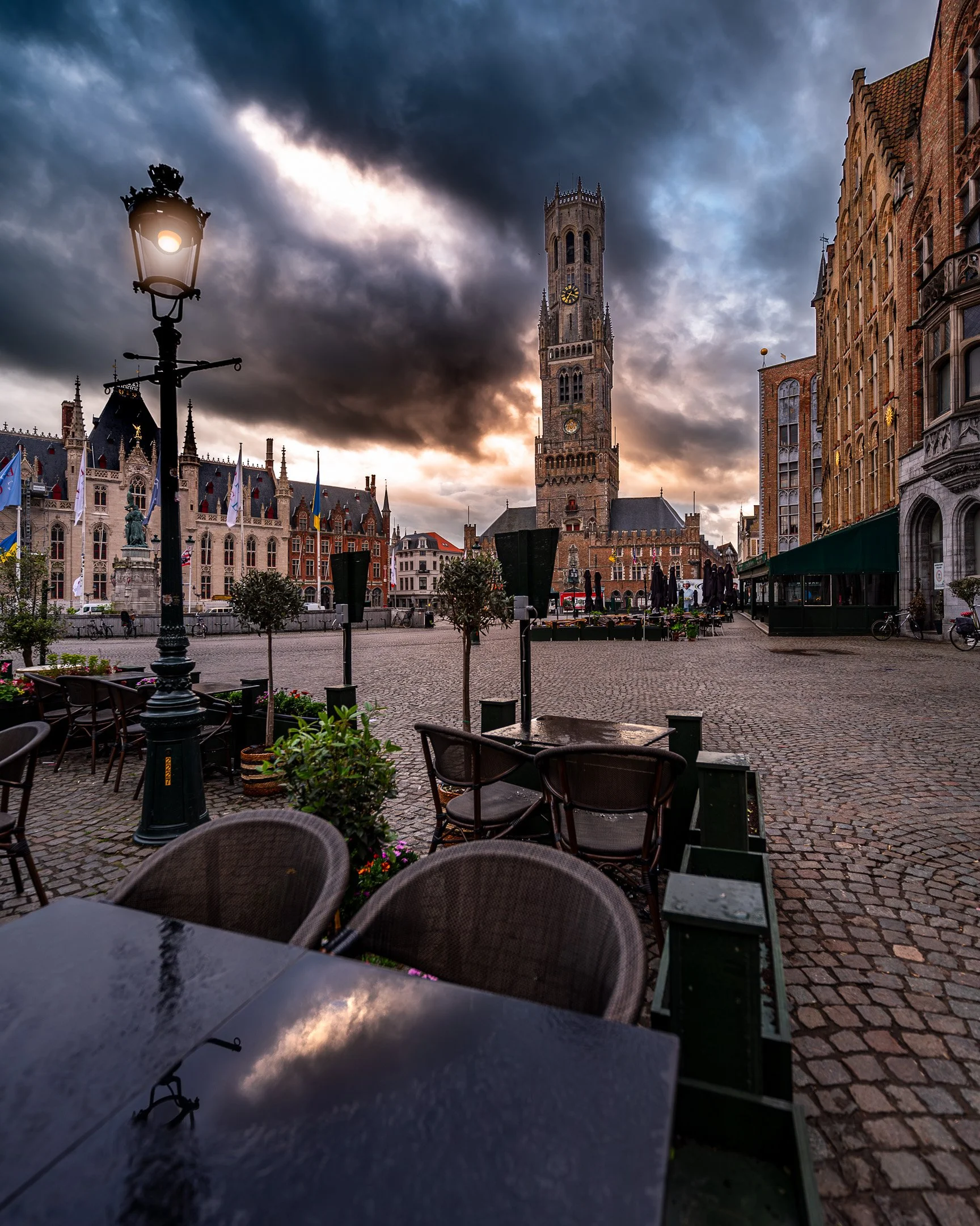 A scenic view of a European town square at sunset, featuring a tall clock tower, old brick buildings, outdoor seating with tables and chairs, street lamps, and a cloudy sky.