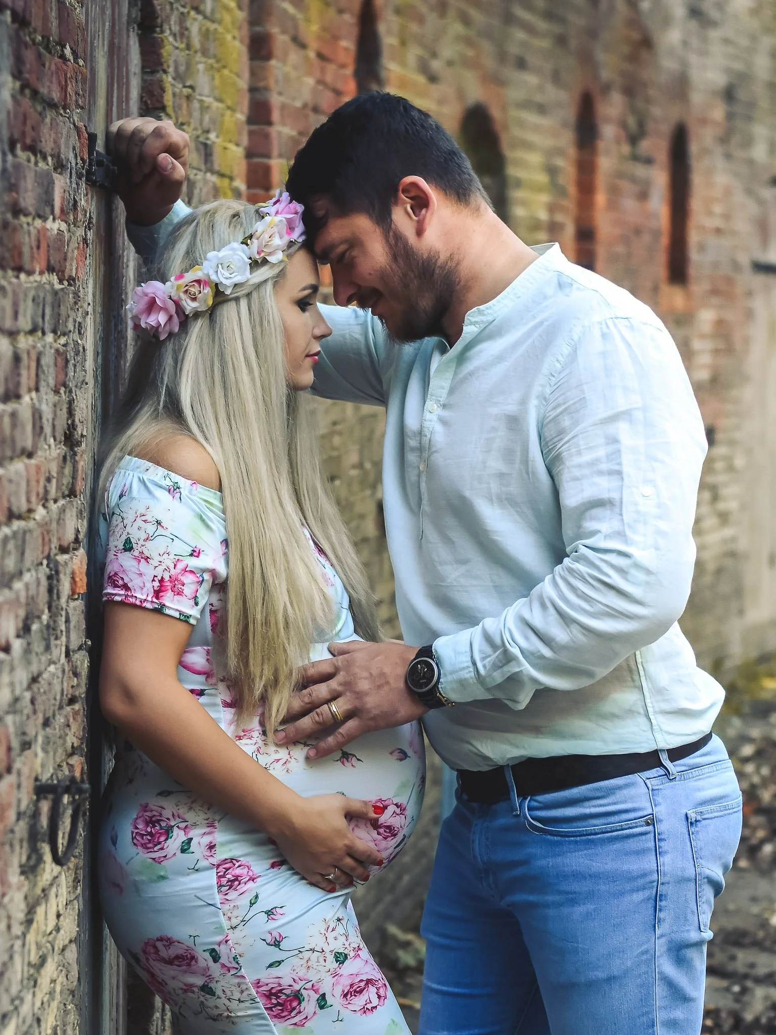 A pregnant woman with long blonde hair wearing a floral dress and flower crown, leaning against a brick wall, facing a man with dark hair and beard wearing a light shirt and jeans, both with foreheads touching in an intimate moment.