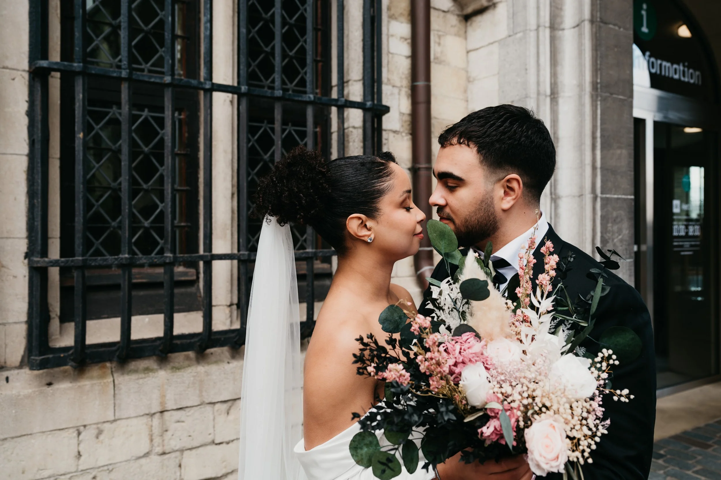 A bride and groom close together, face to face, outside a building with beige stone walls and black iron window bars, with the bride holding a large bouquet of pink and white flowers.