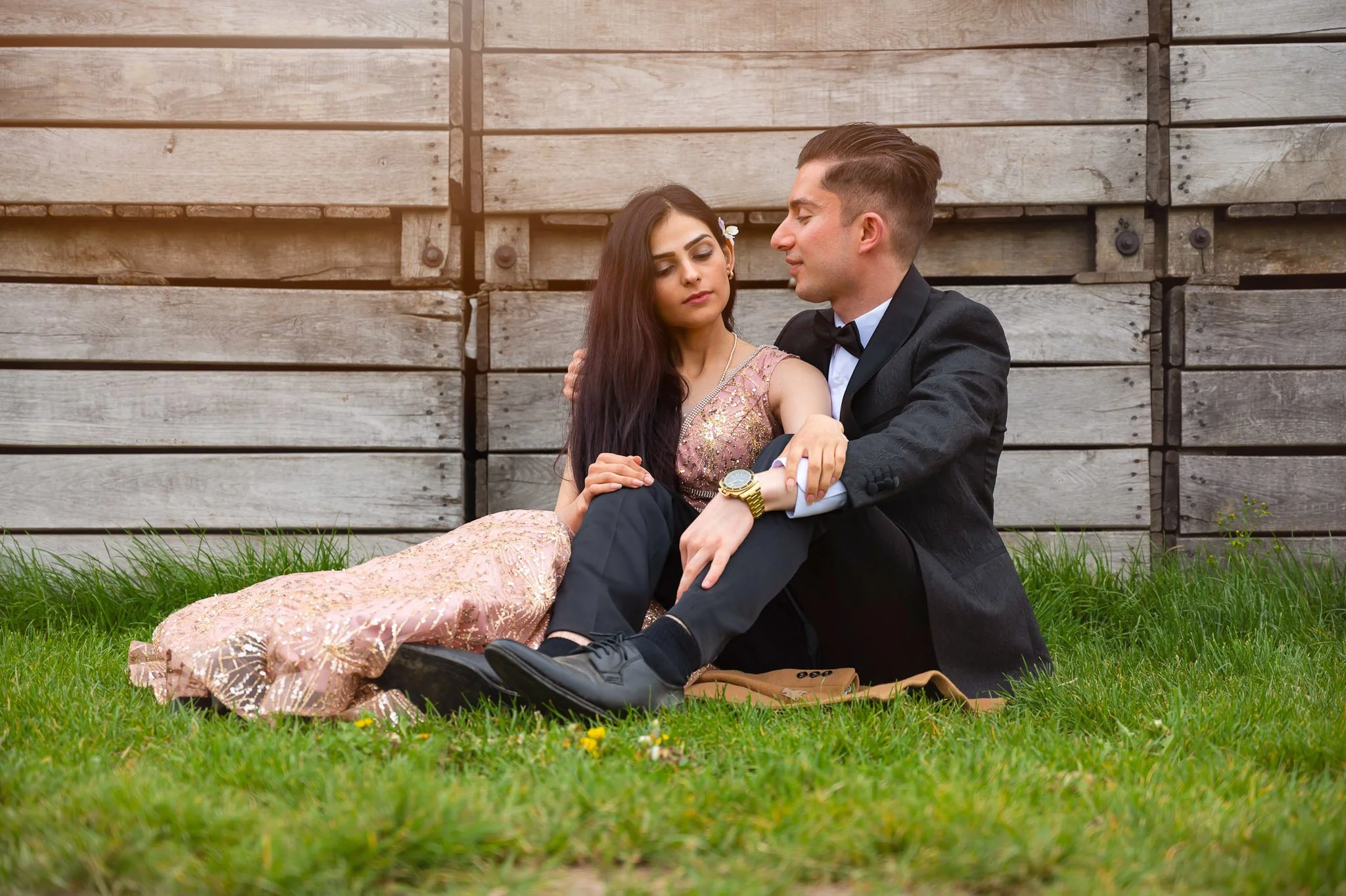 A young man and woman sitting on grass near a wooden fence, dressed in formal attire, with the man wearing a tuxedo and the woman in a pink gown, sharing an intimate moment.