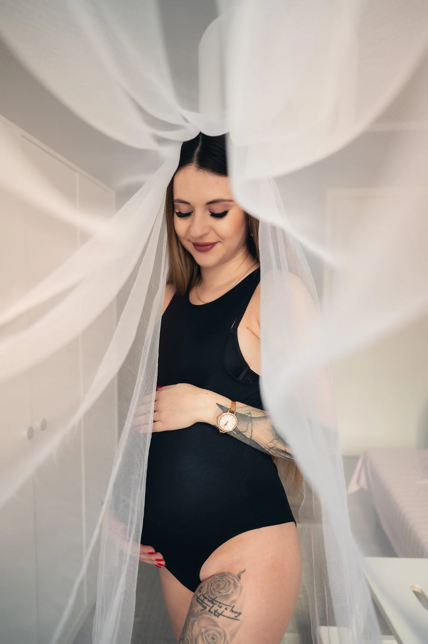 A young woman with long brown hair, tattoos on her left arm and thigh, wearing a black sleeveless top and underwear, is standing behind a sheer white curtain in a room with white walls and a bed.