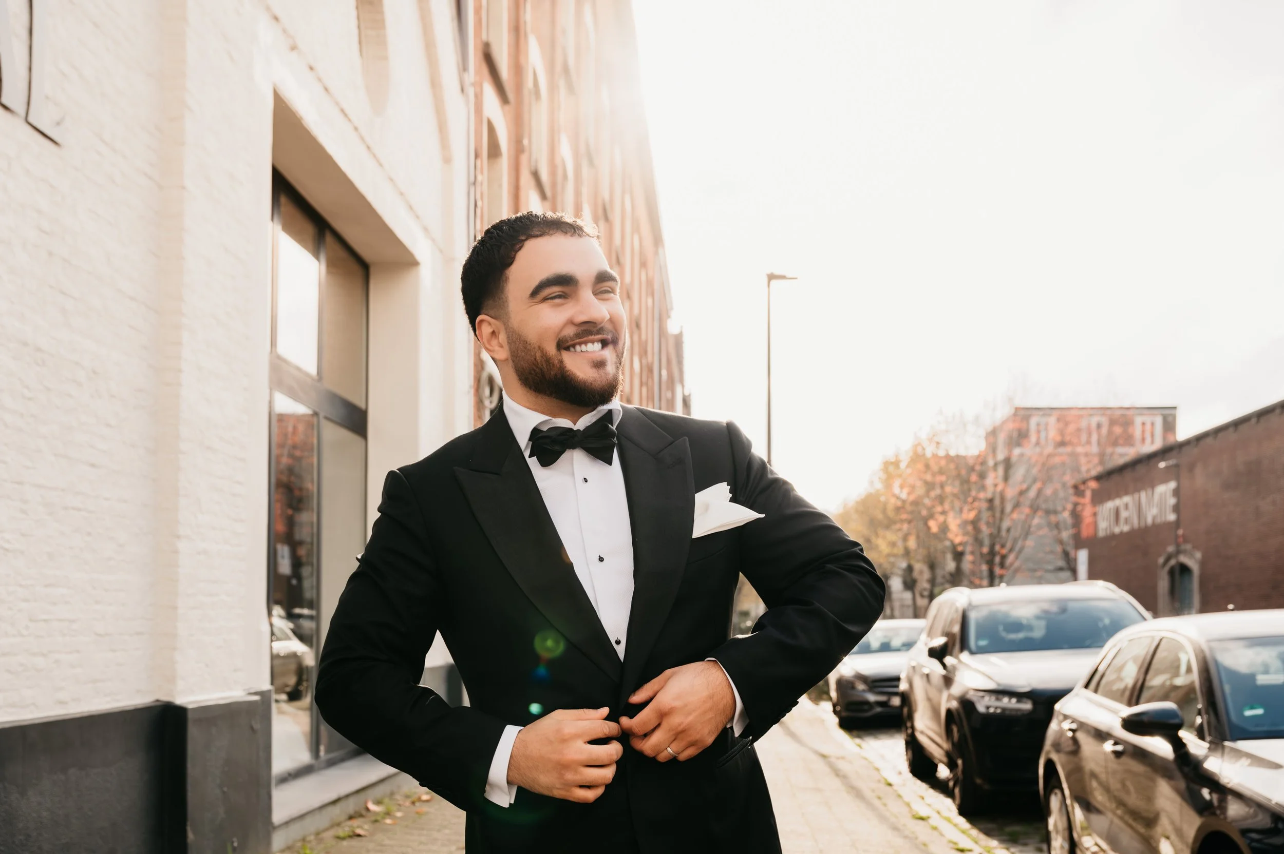 A man in a black tuxedo and bow tie smiling while adjusting his jacket on a city sidewalk in the late afternoon.