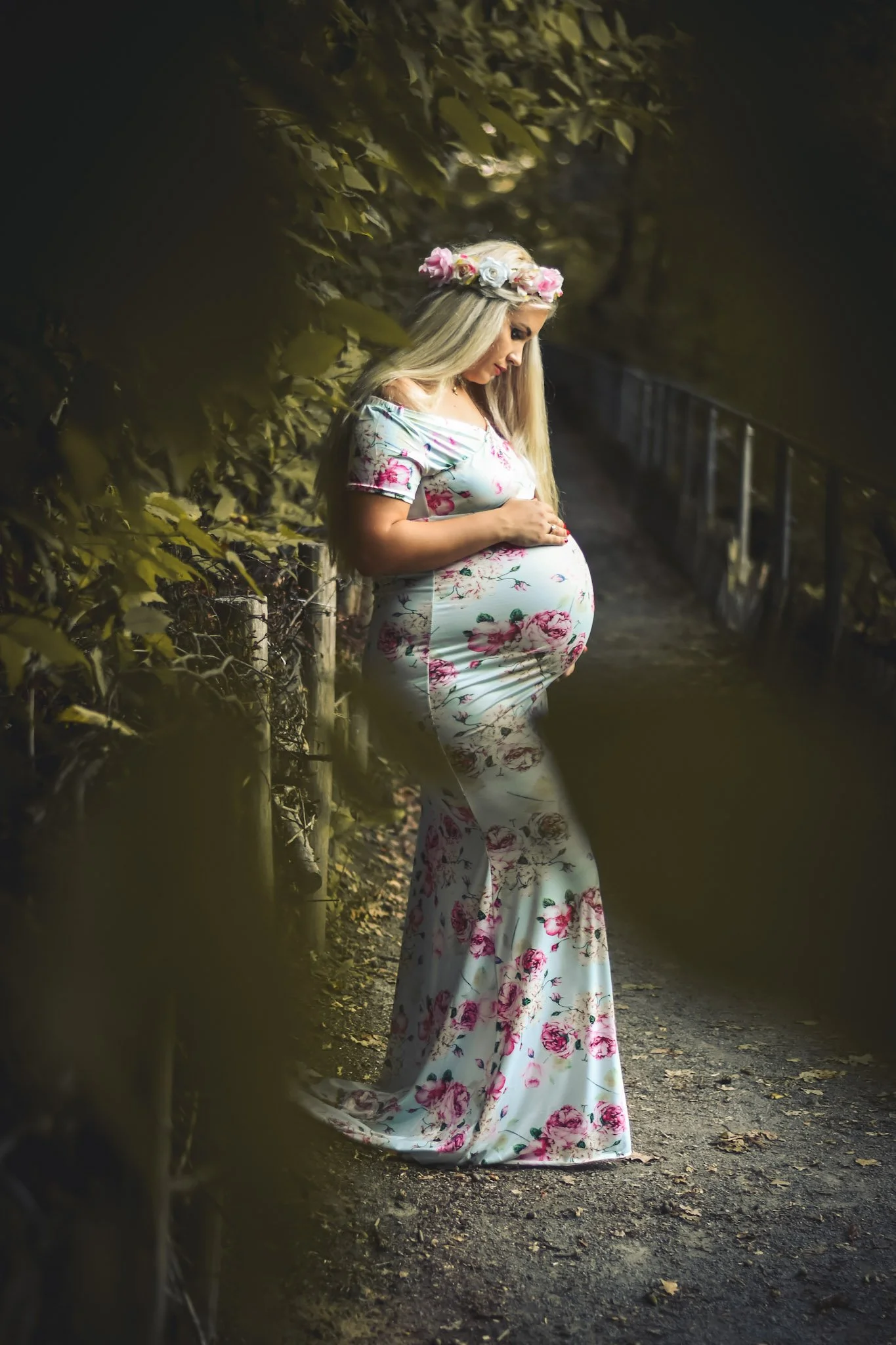 Pregnant woman wearing a floral dress and flower crown, standing on a forest path, looking down and gently holding her belly.