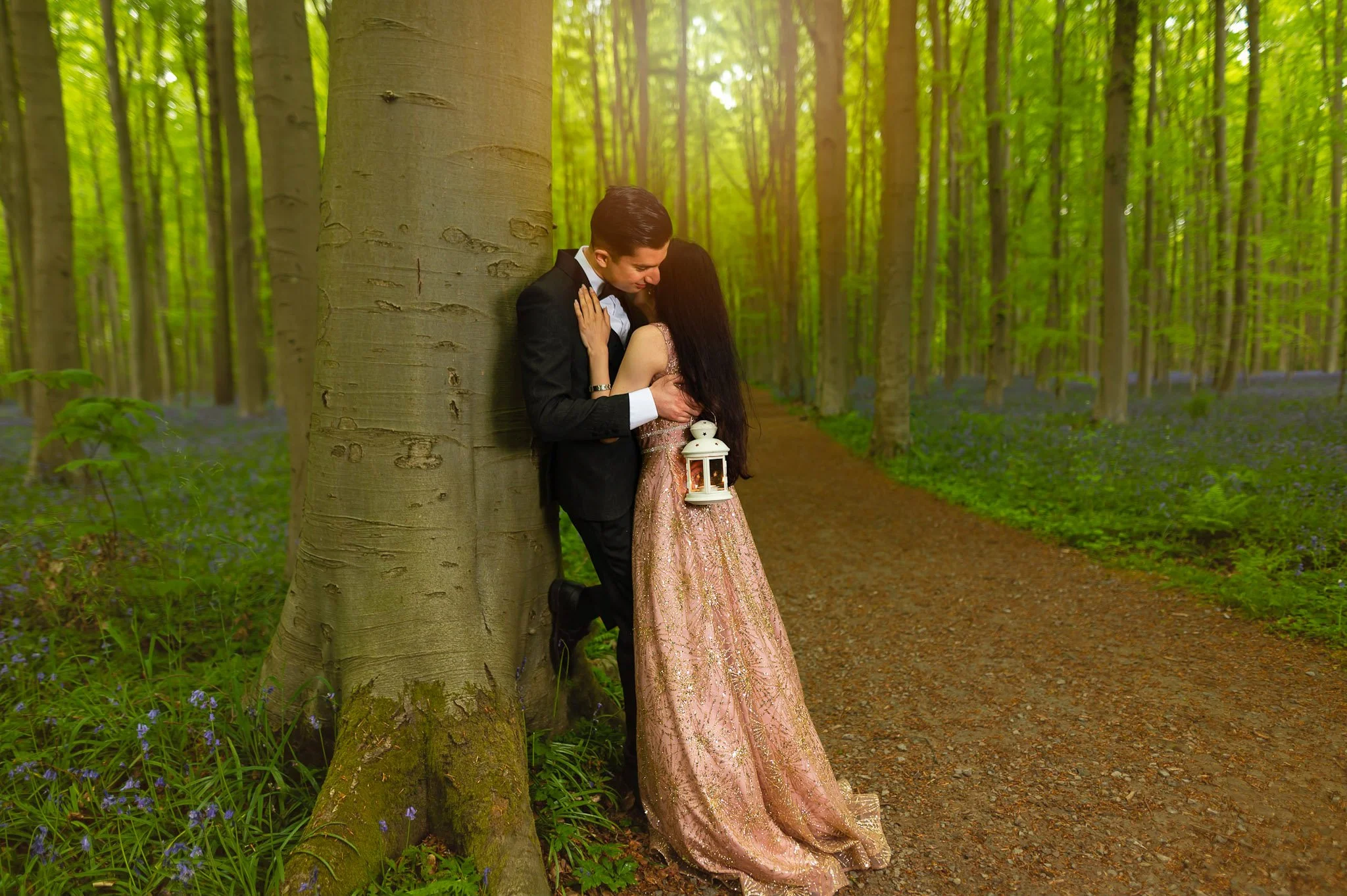 A romantic couple in formal attire embracing and smiling in a lush green forest with a dirt path, during daytime.