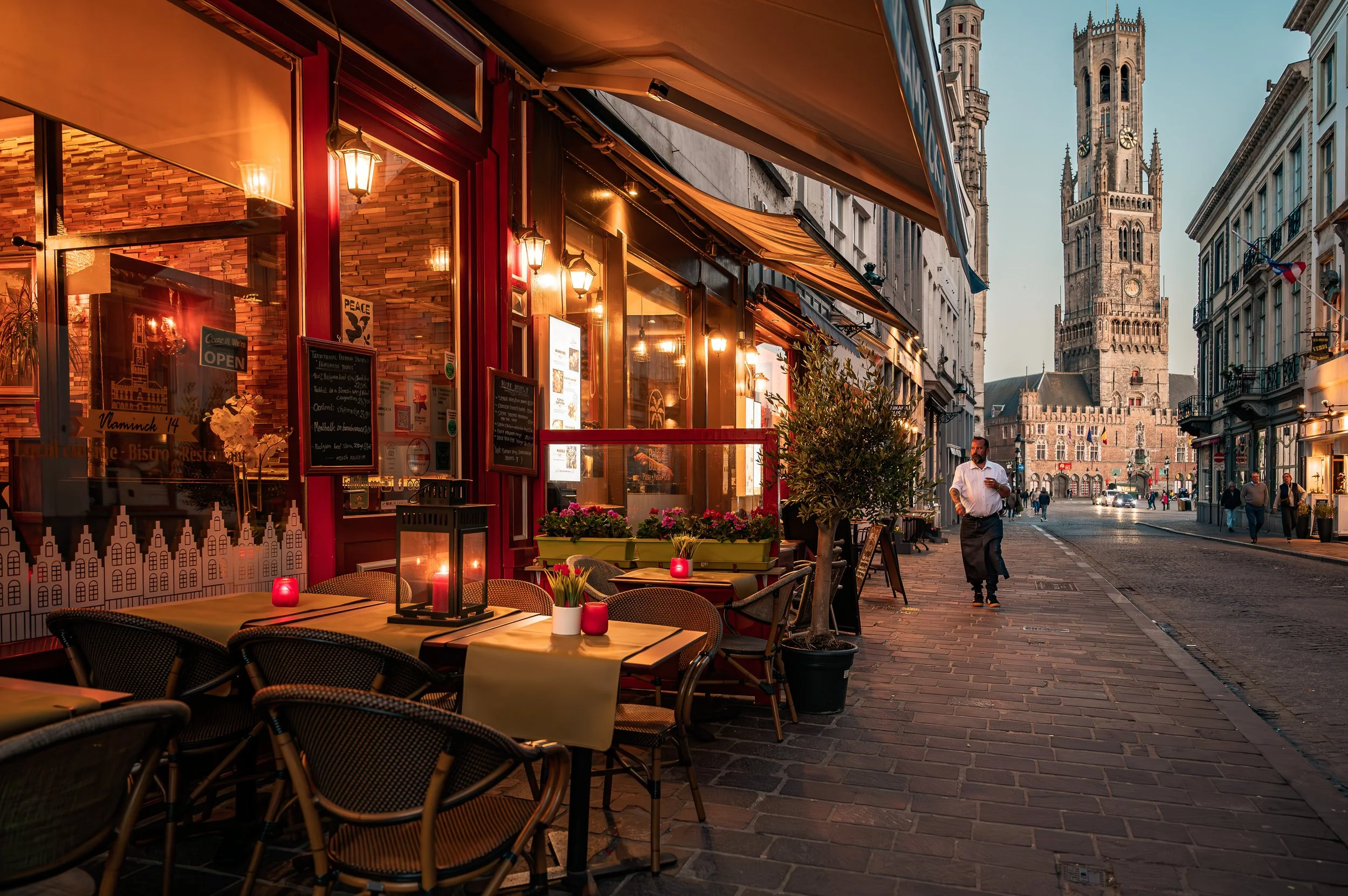 An outdoor café along a cobblestone street with tables decorated with candles and flowers, near a historic clock tower in a European city at dusk.