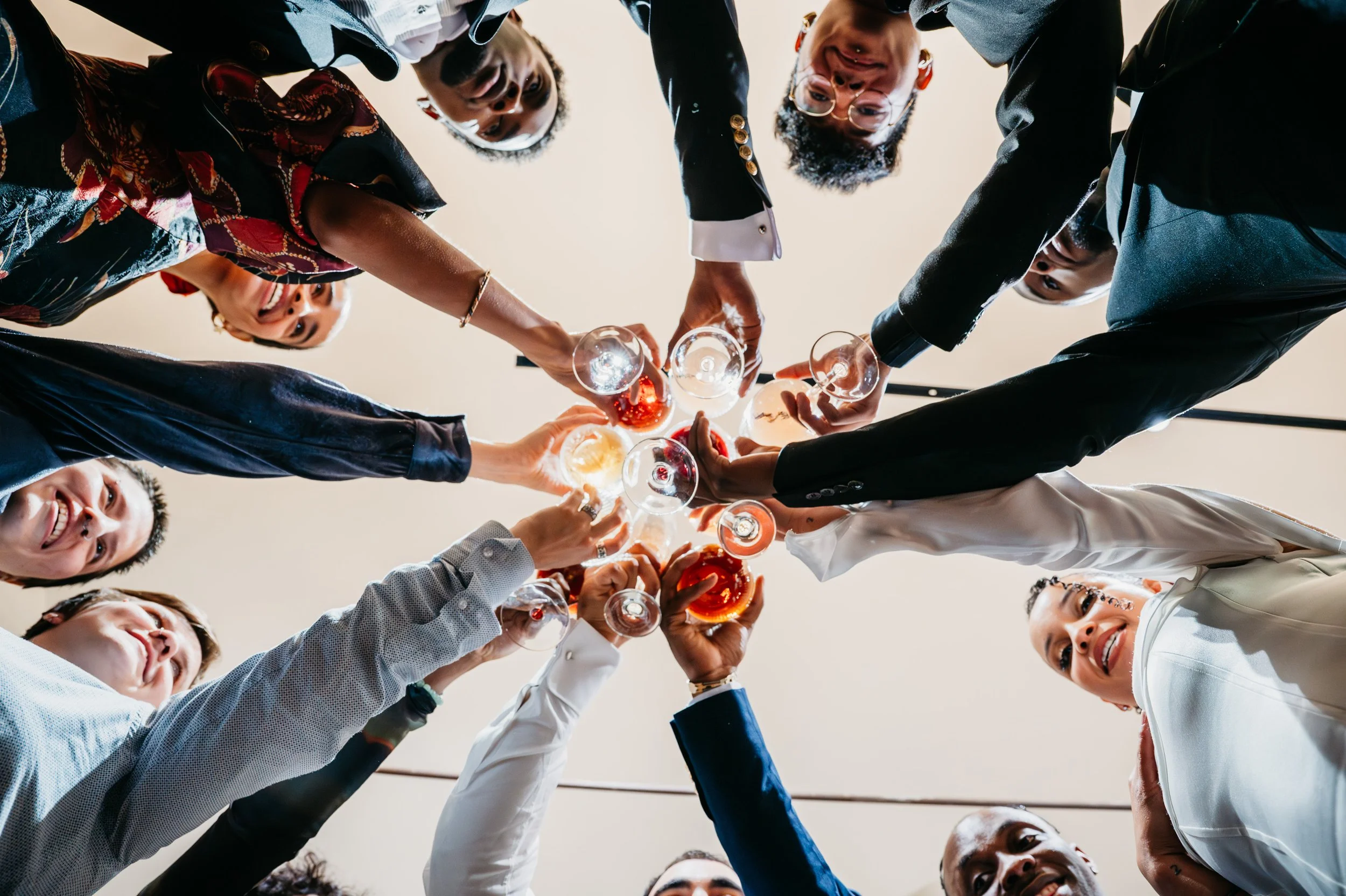 A diverse group of people in formal attire cheers together with glasses of wine or champagne raised in a circle, viewed from below.