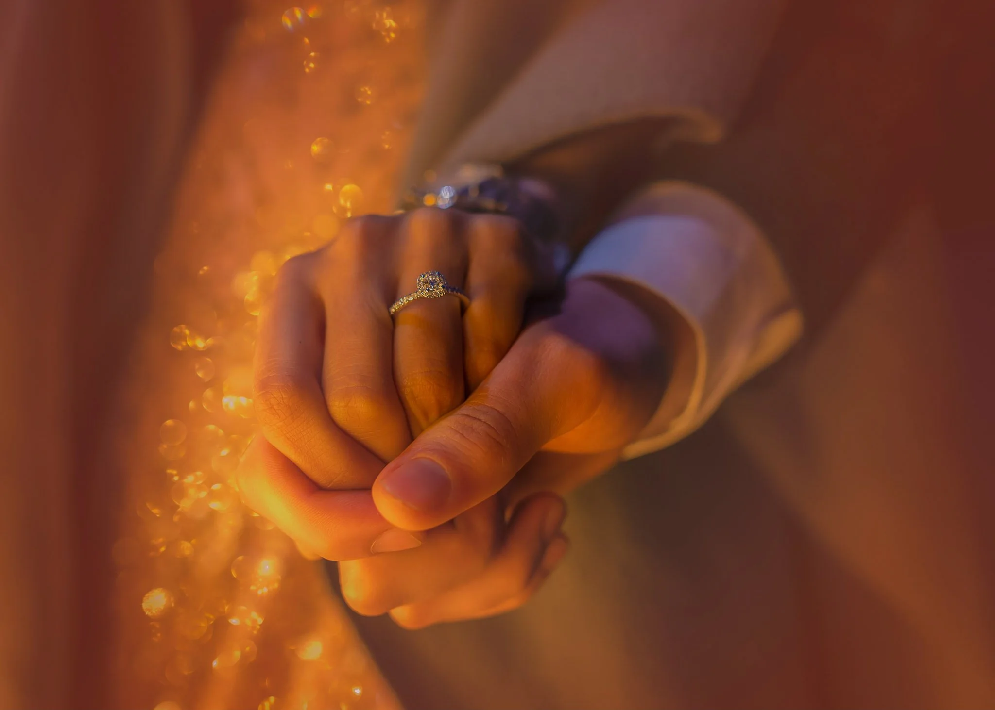Close-up of a person's hand wearing a diamond engagement ring, resting on a golden, glittery surface, with a satin fabric sleeve partially visible.