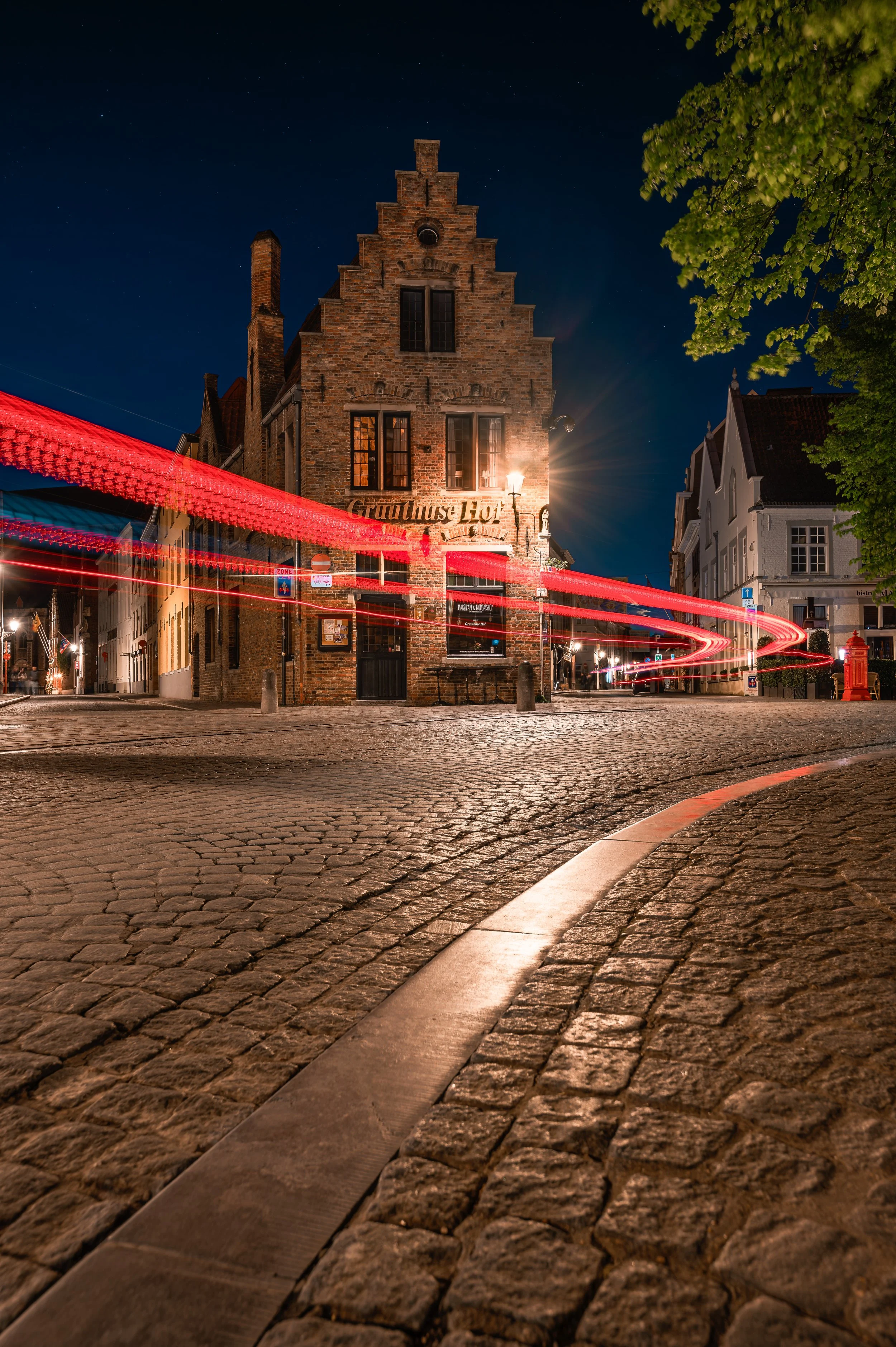 Nighttime scene of a cobblestone street with a historic brick building labeled "Gruithuys 1516" in the center, light trails from passing vehicles, a large green-leafed tree on the right, and a starry sky overhead.