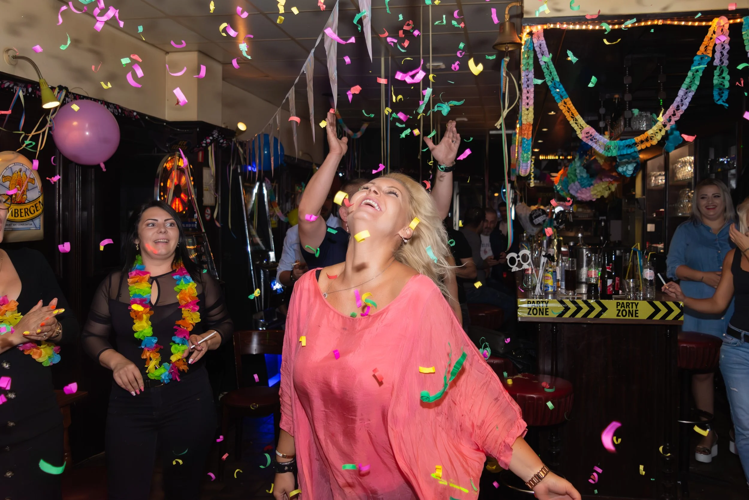 A group of women celebrating at a party, with colorful confetti falling, balloons, and decorations, in a lively indoor setting.