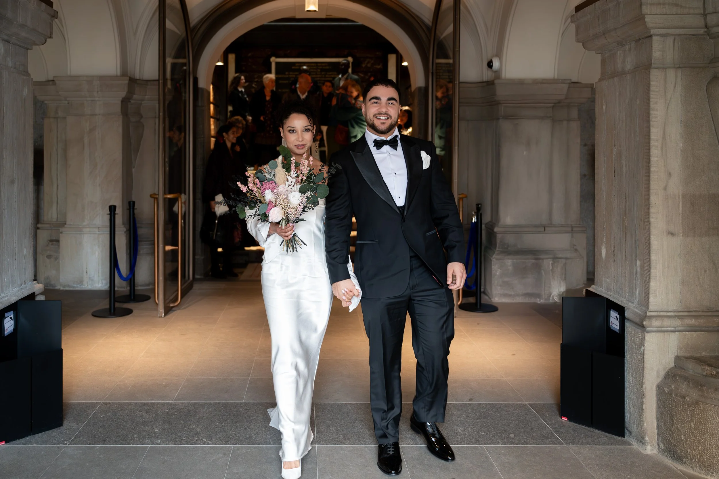 A newlywed couple walking hand-in-hand out of a church, with the bride holding a large bouquet of flowers, both smiling and dressed in wedding attire.