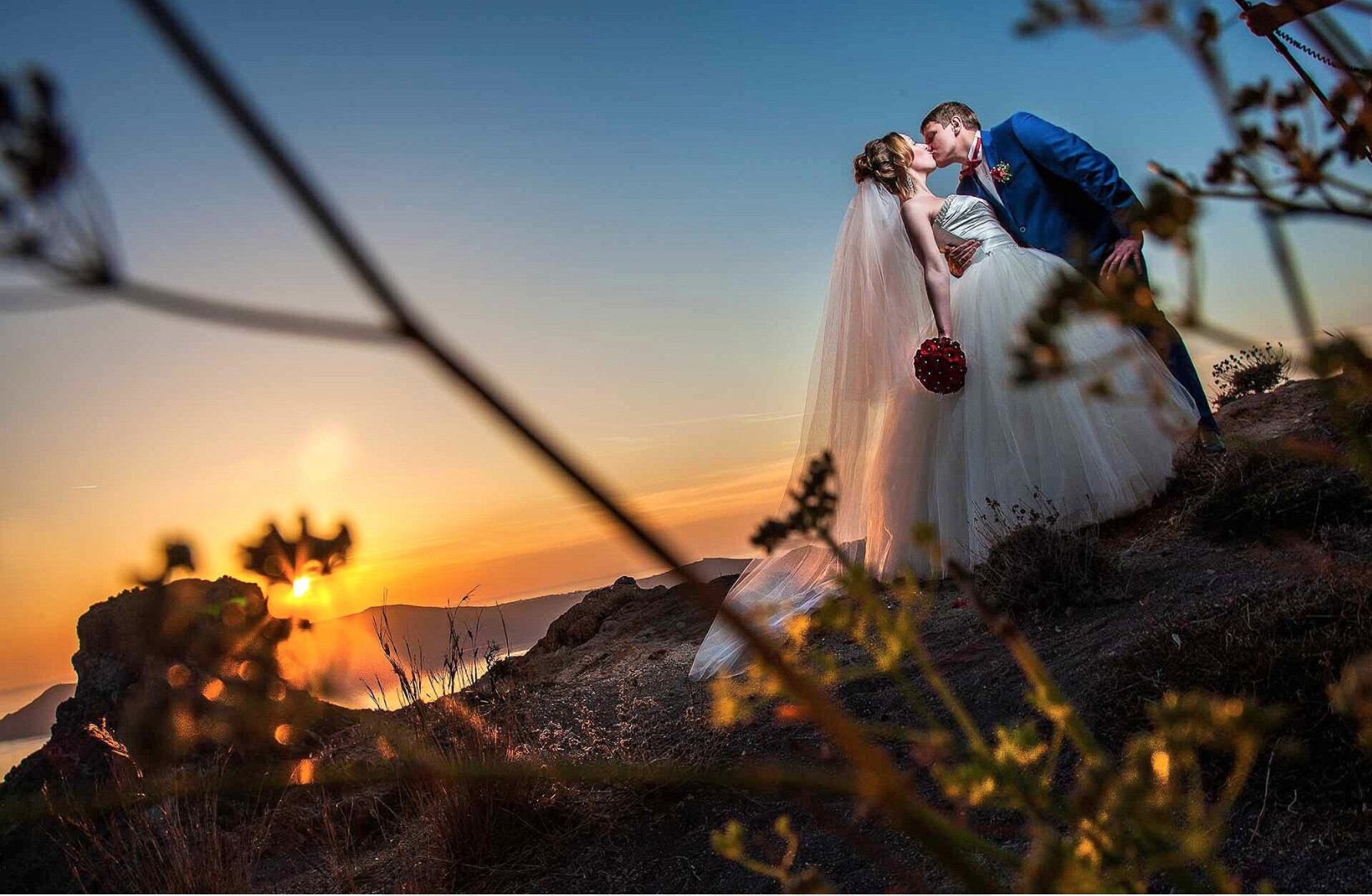 Iconic wide shot of a couple near Oia’s blue domes with panoramic caldera backdrop, capturing classic Santorini wedding imagery at golden hour.