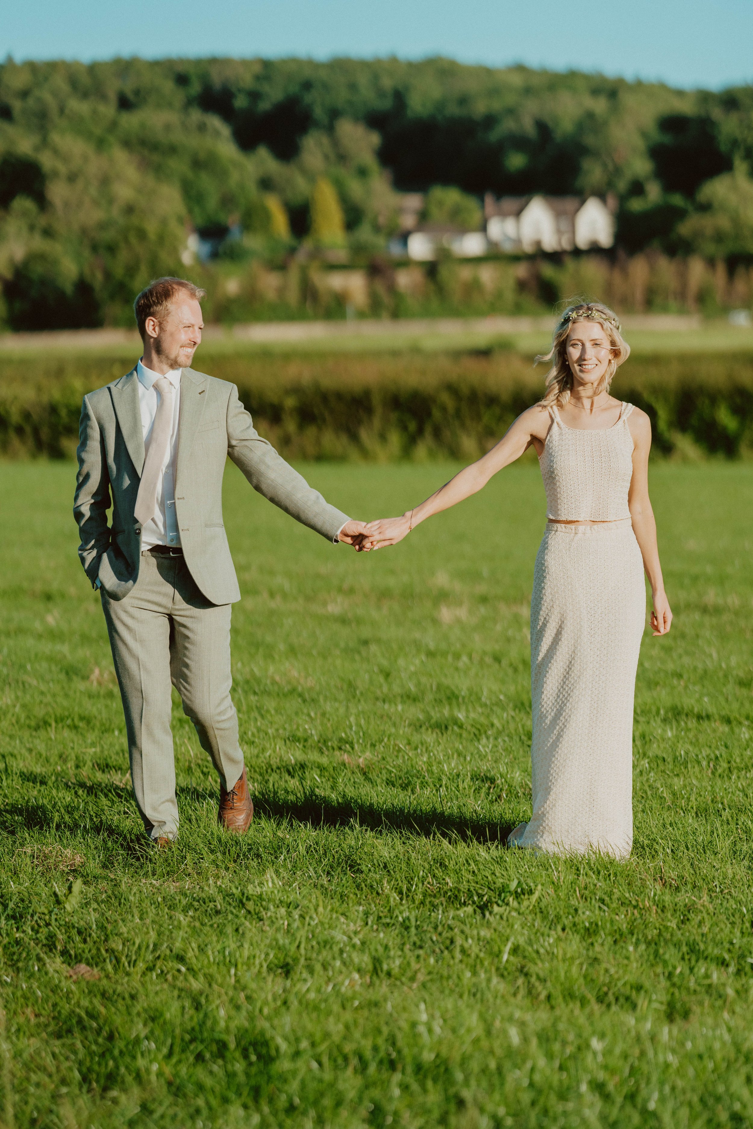 A man in a light gray suit and a woman in a cream dress holding hands outdoors on a green field during daylight with a lake and hills in the background.