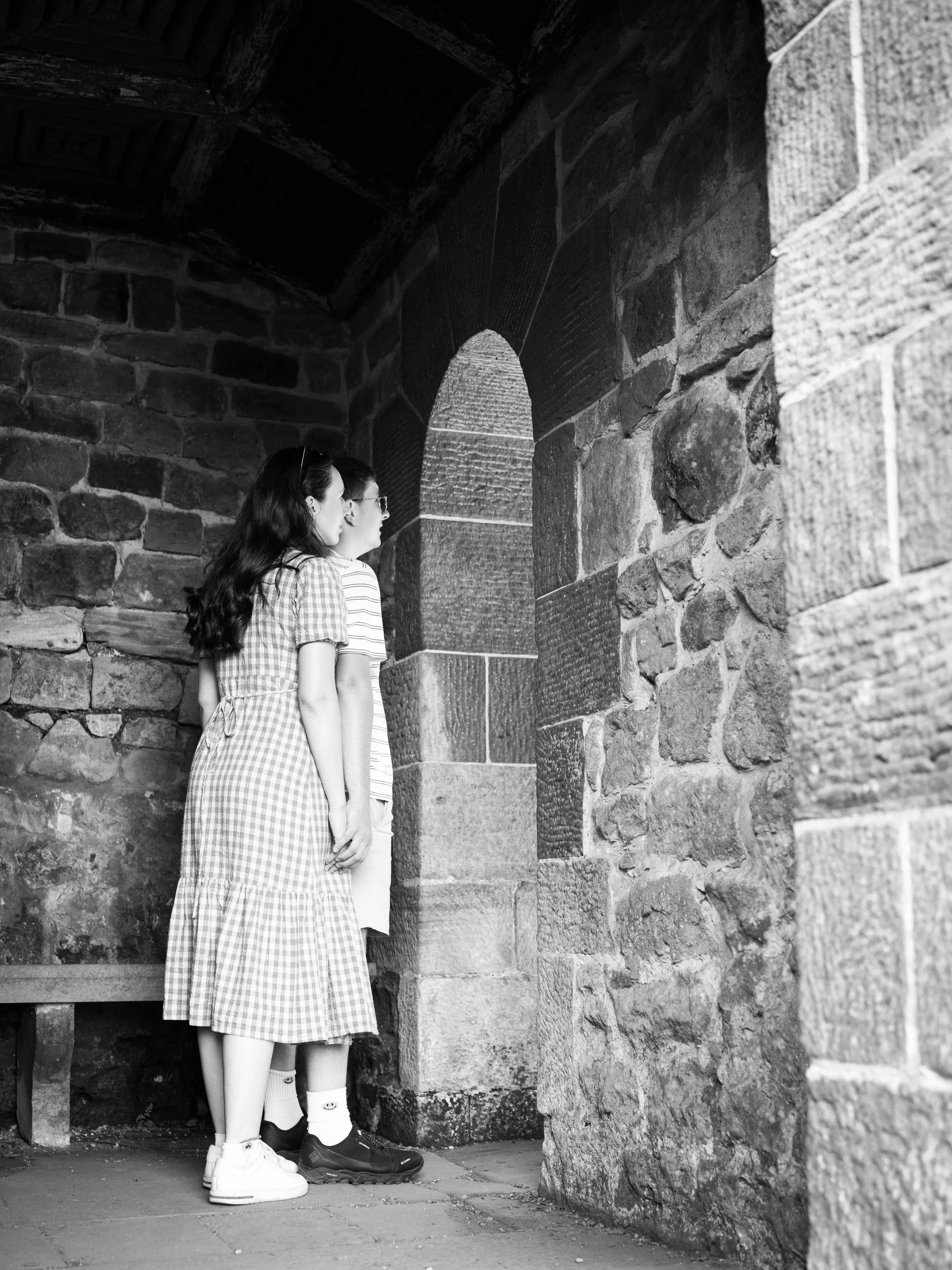 A black and white photo of a boy and girl standing inside a stone structure, looking through a narrow arched window.