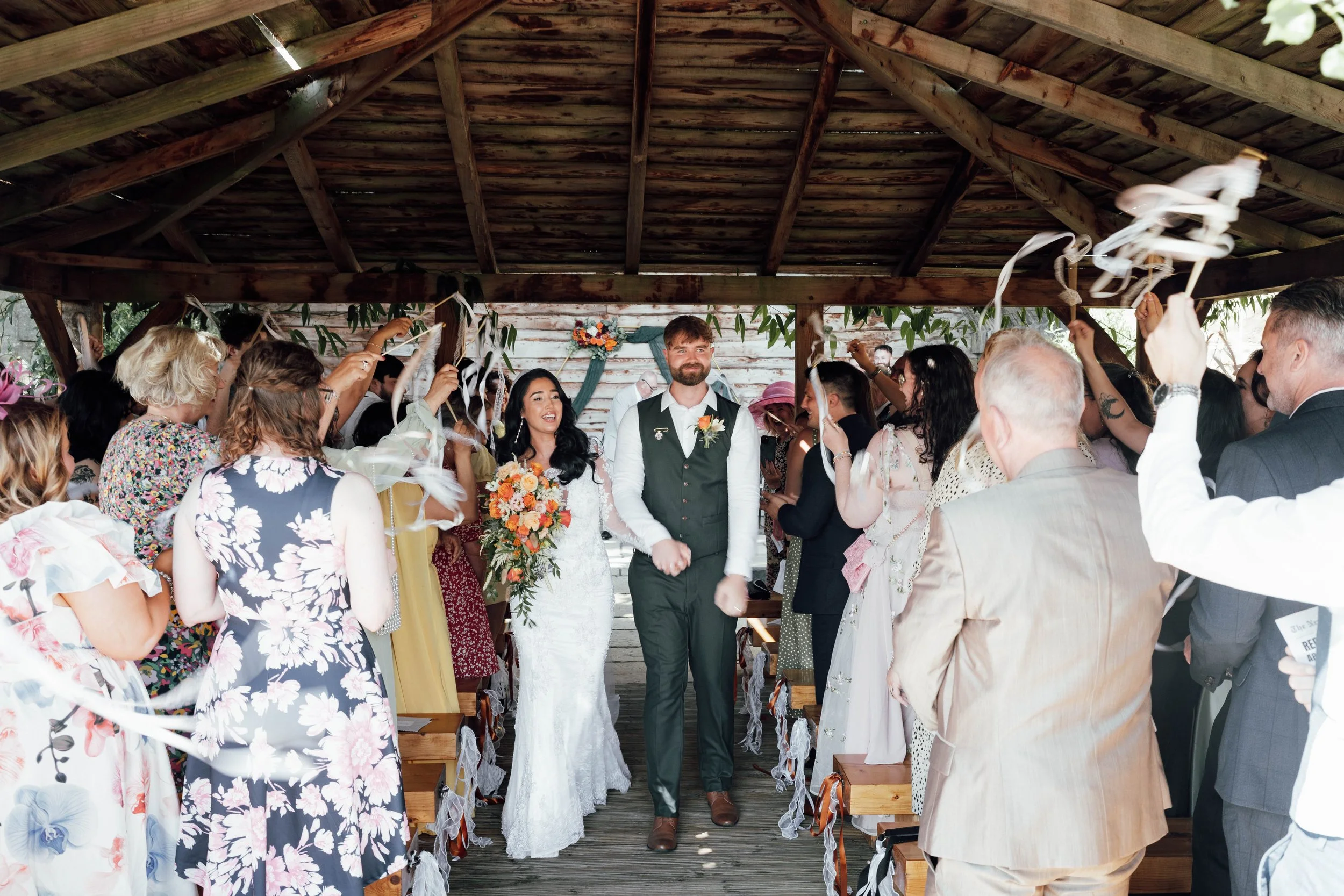 A wedding ceremony with a bride, groom, and guests under a wooden pavilion. The bride holds a bouquet, and guests are holding streamers.