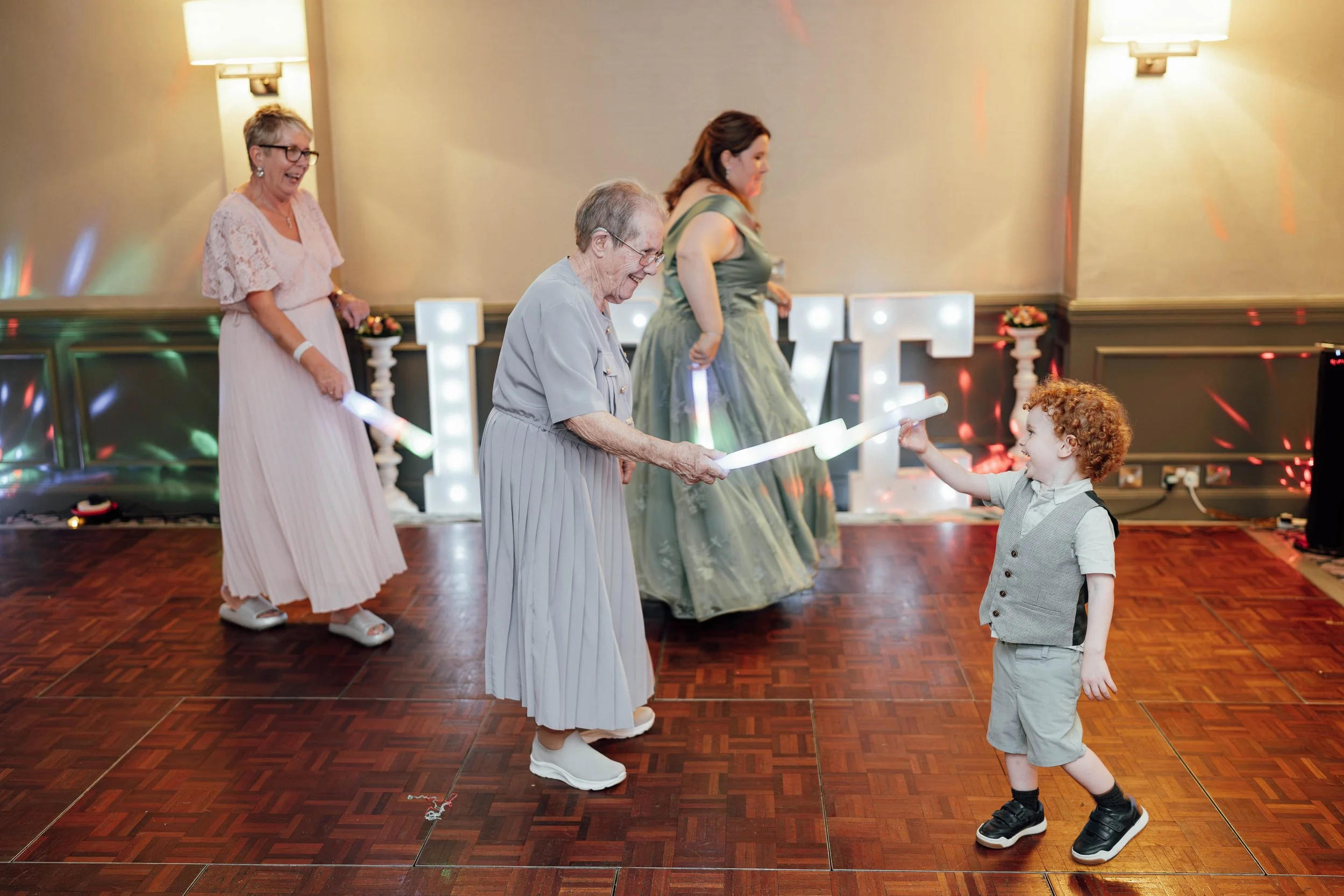 Four women and a young boy with red curly hair dancing and playing with illuminated batons on a wooden dance floor at a celebration event.