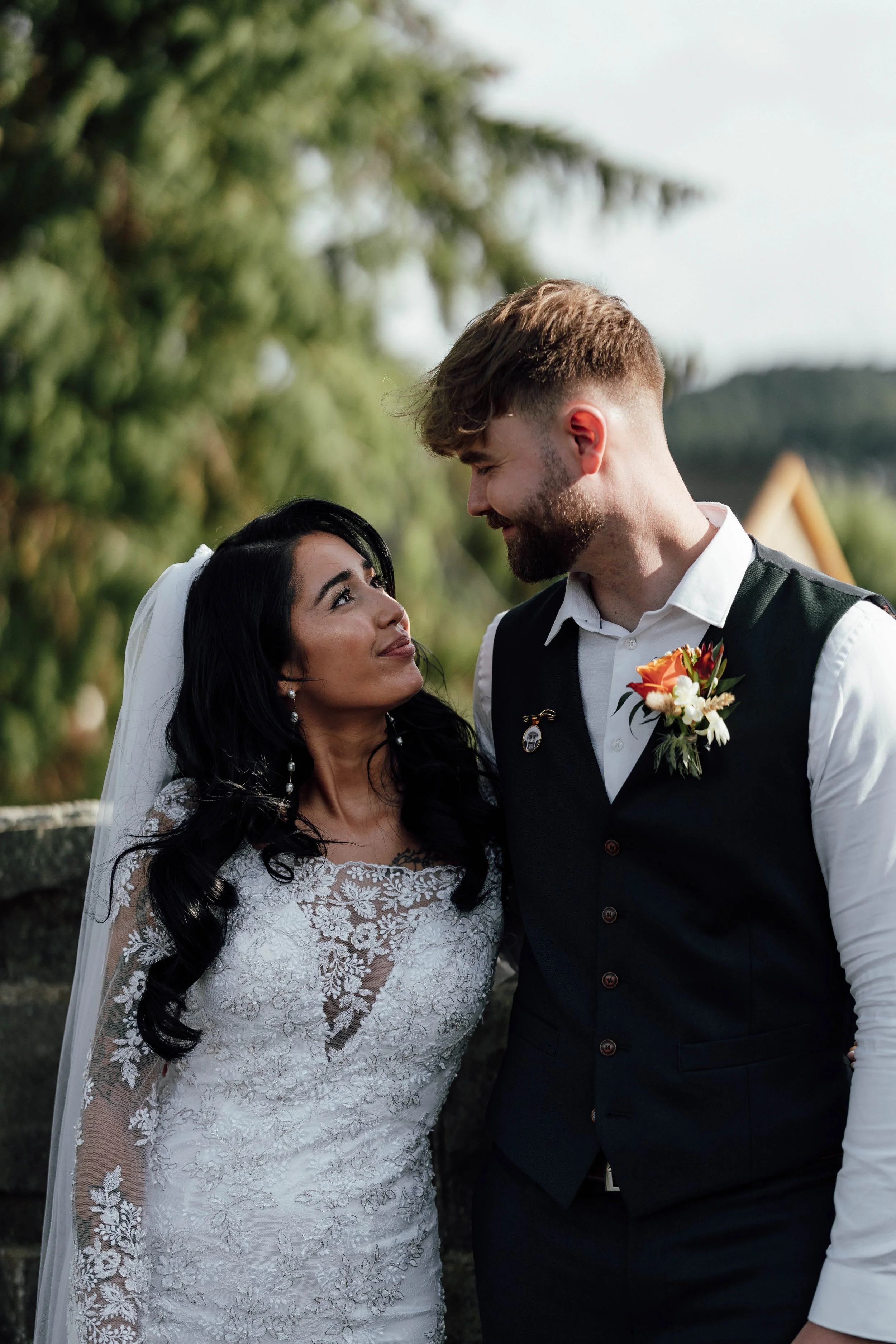 A bride and groom sharing a loving moment outdoors on their wedding day, looking into each other's eyes with trees and a house in the background.