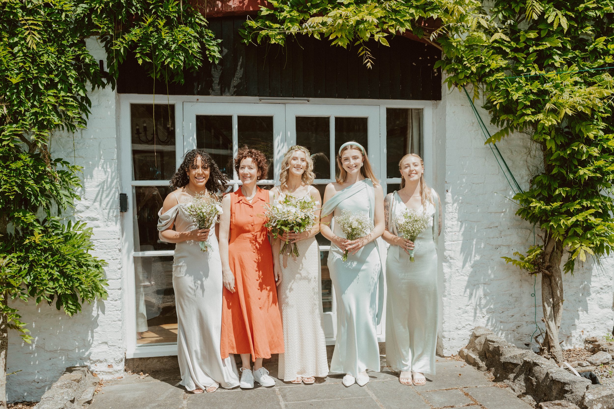 Five women standing outside in front of a white brick building with greenery, wearing dresses, holding bouquets, smiling for a photo.