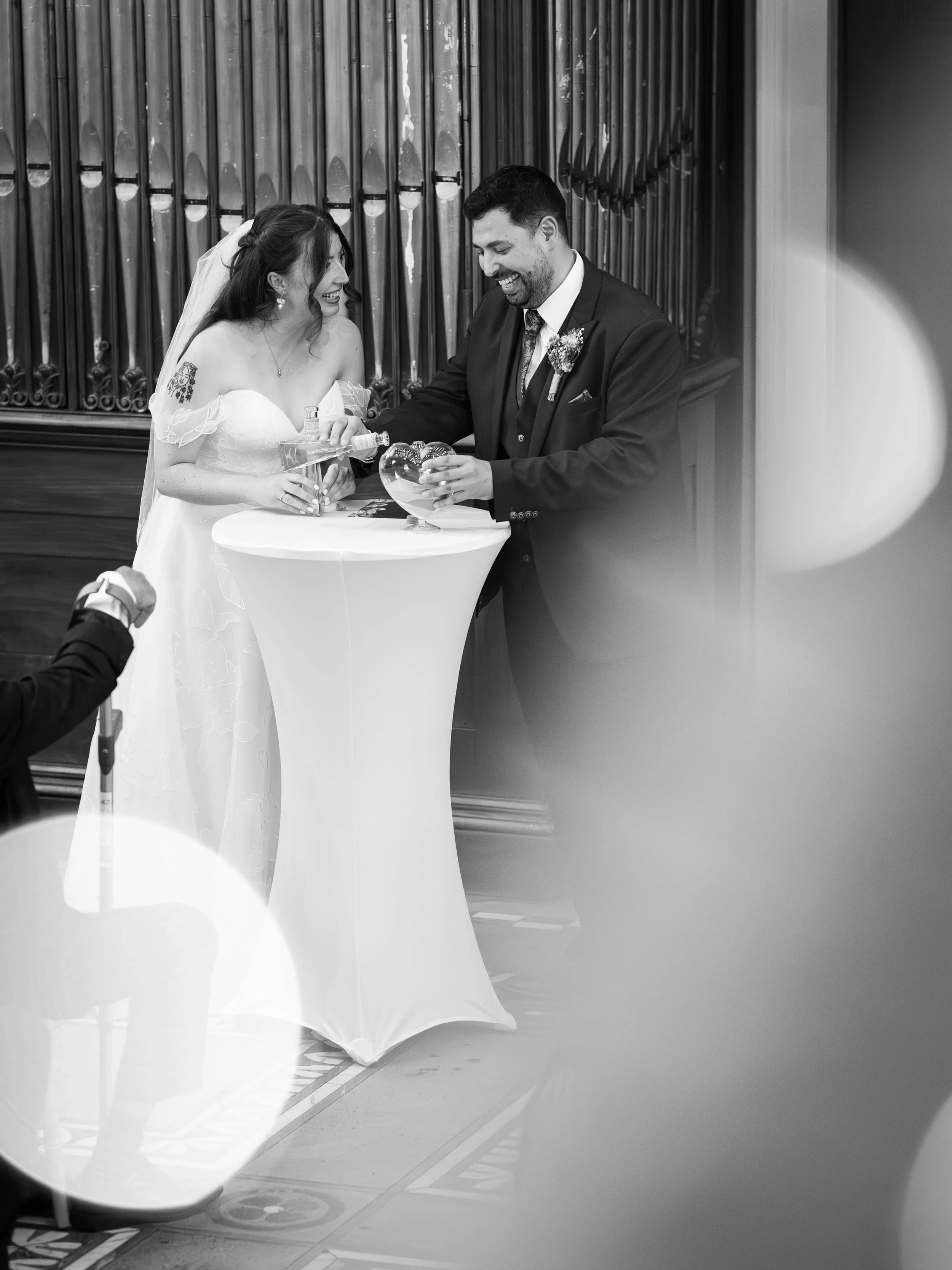 A bride and groom ring the wedding ceremony, smiling and sharing a moment during their vows at a small table, with a background of pipe organ pipes.