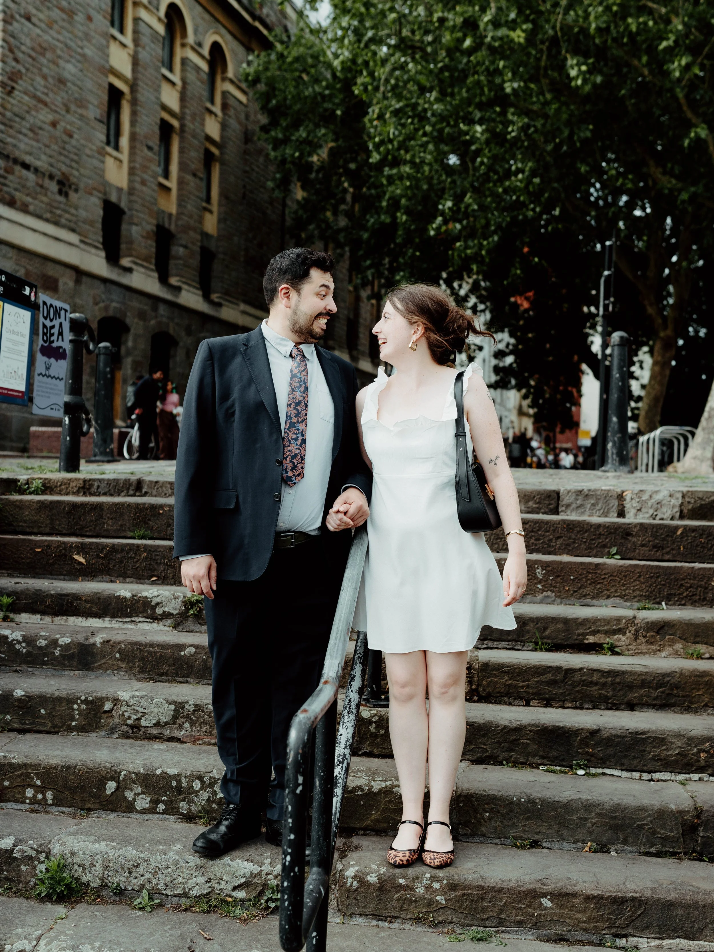 A man in a suit and a woman in a white dress hold hands and smile at each other on a set of steps outdoors.