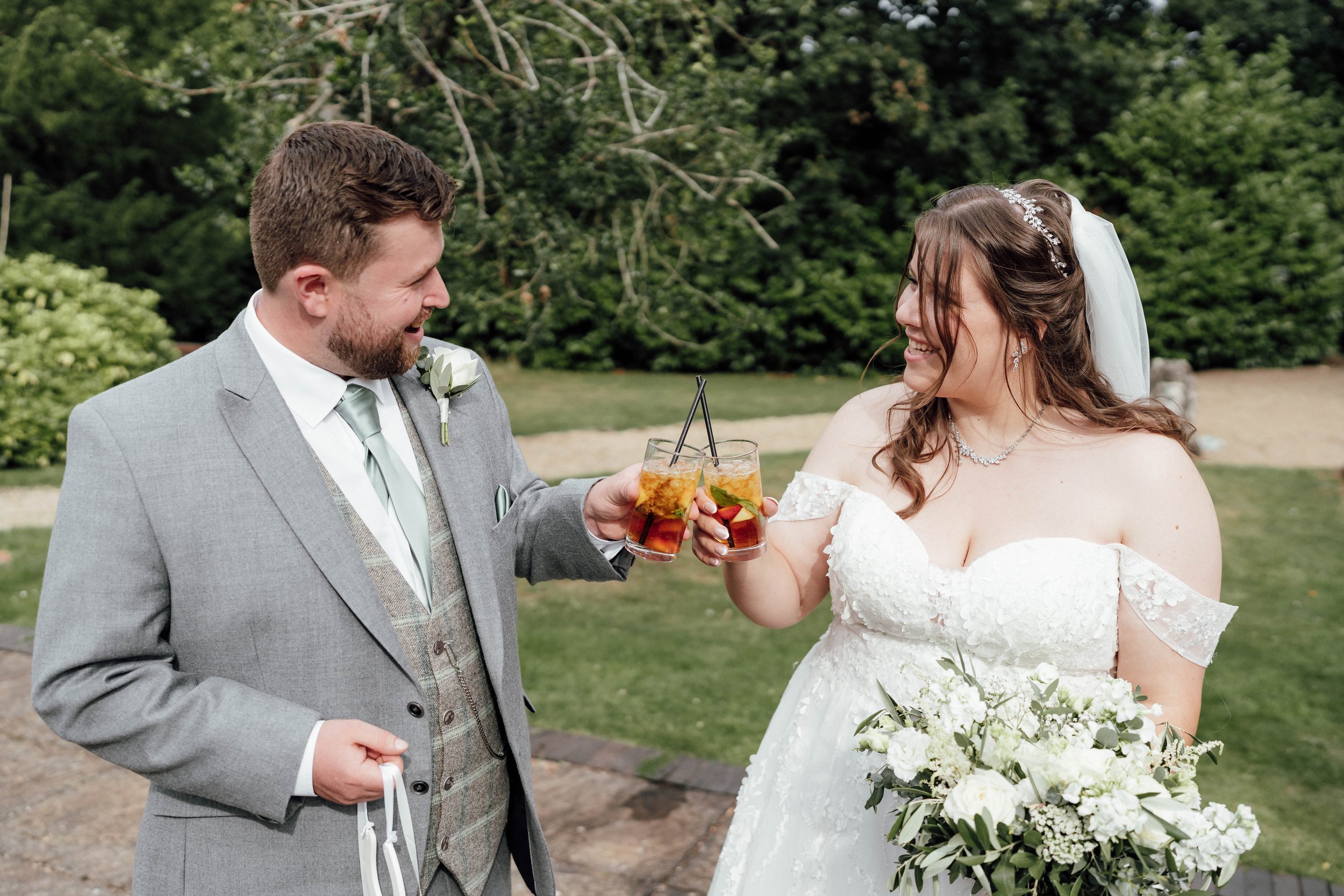 A newlywed couple in wedding attire raising drinks in a toast outdoors, with a green garden background.