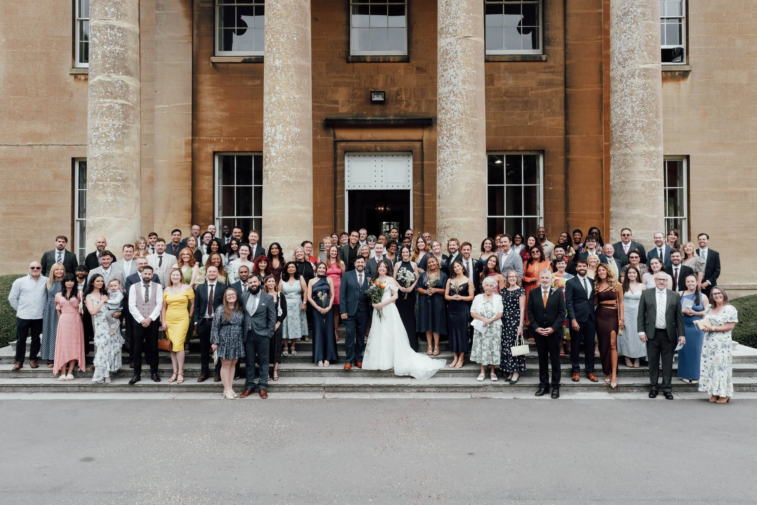 A large group of people in formal attire standing on the steps of a building with tall columns, posing for a photo at a wedding.