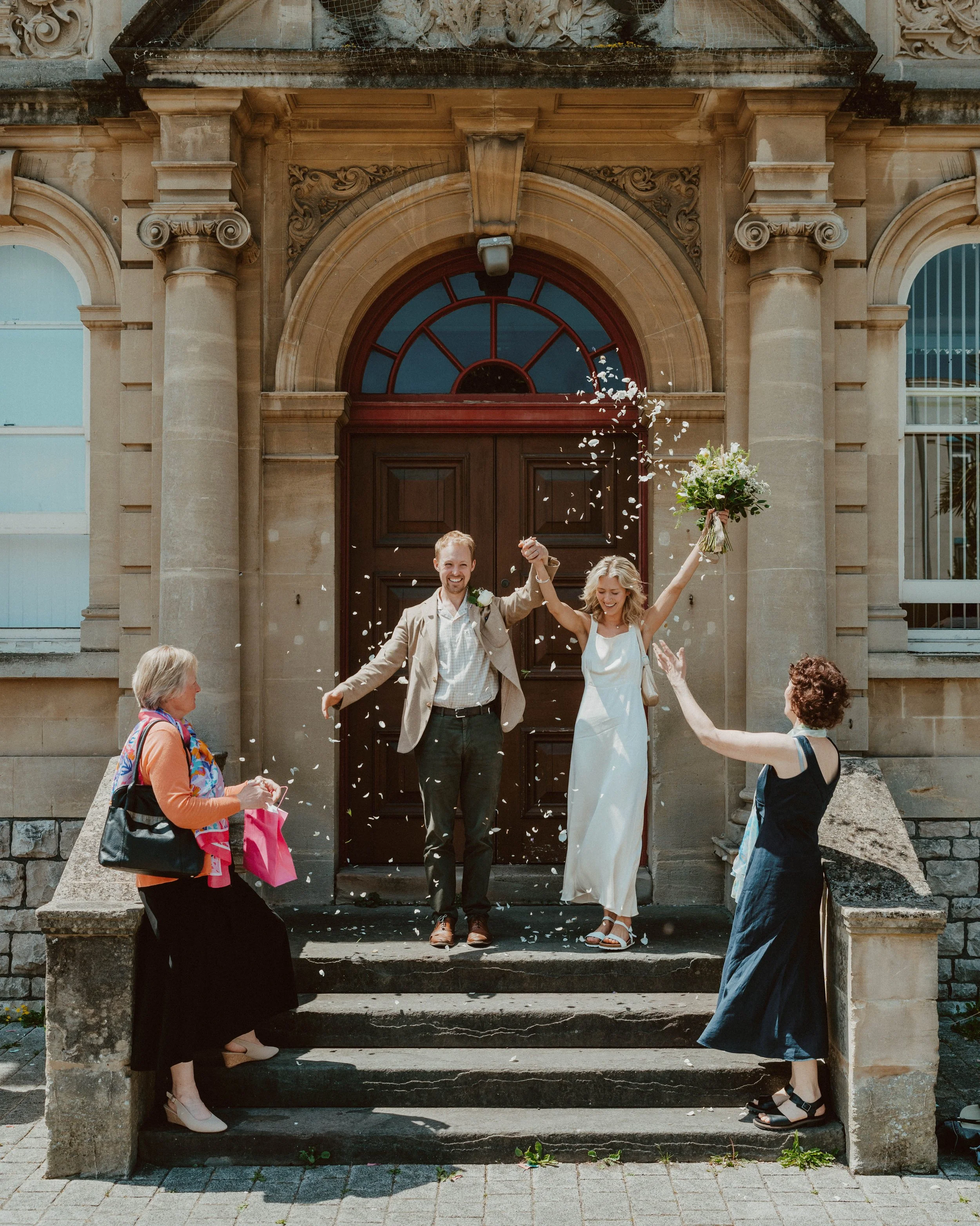 A newlywed couple celebrating on the steps of a church, holding hands with their arms raised, surrounded by family and friends throwing flower petals.