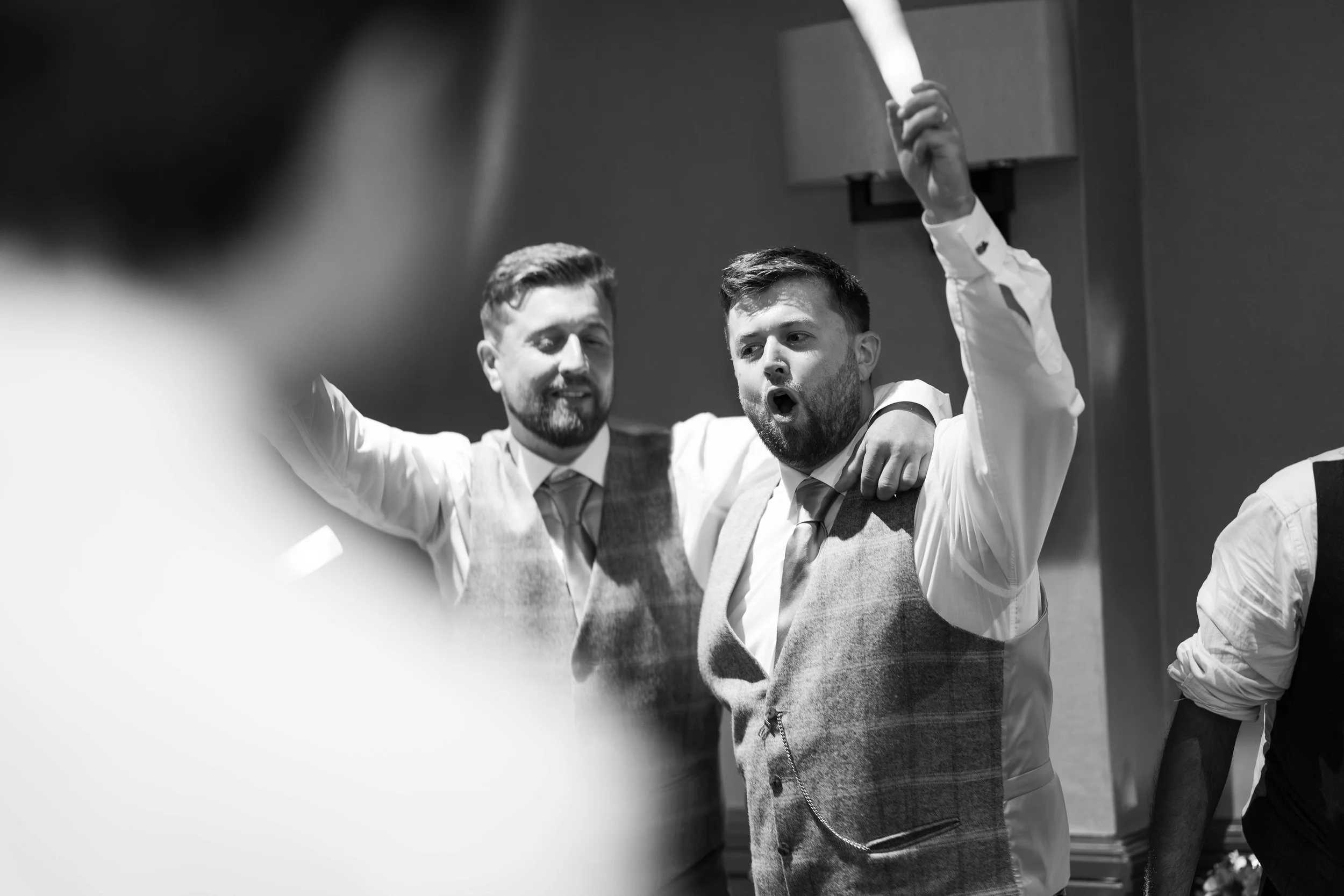 Two men in dress shirts and vests at a celebration, one with arm around the other's shoulder, raising his hand, appearing to sing or cheer.