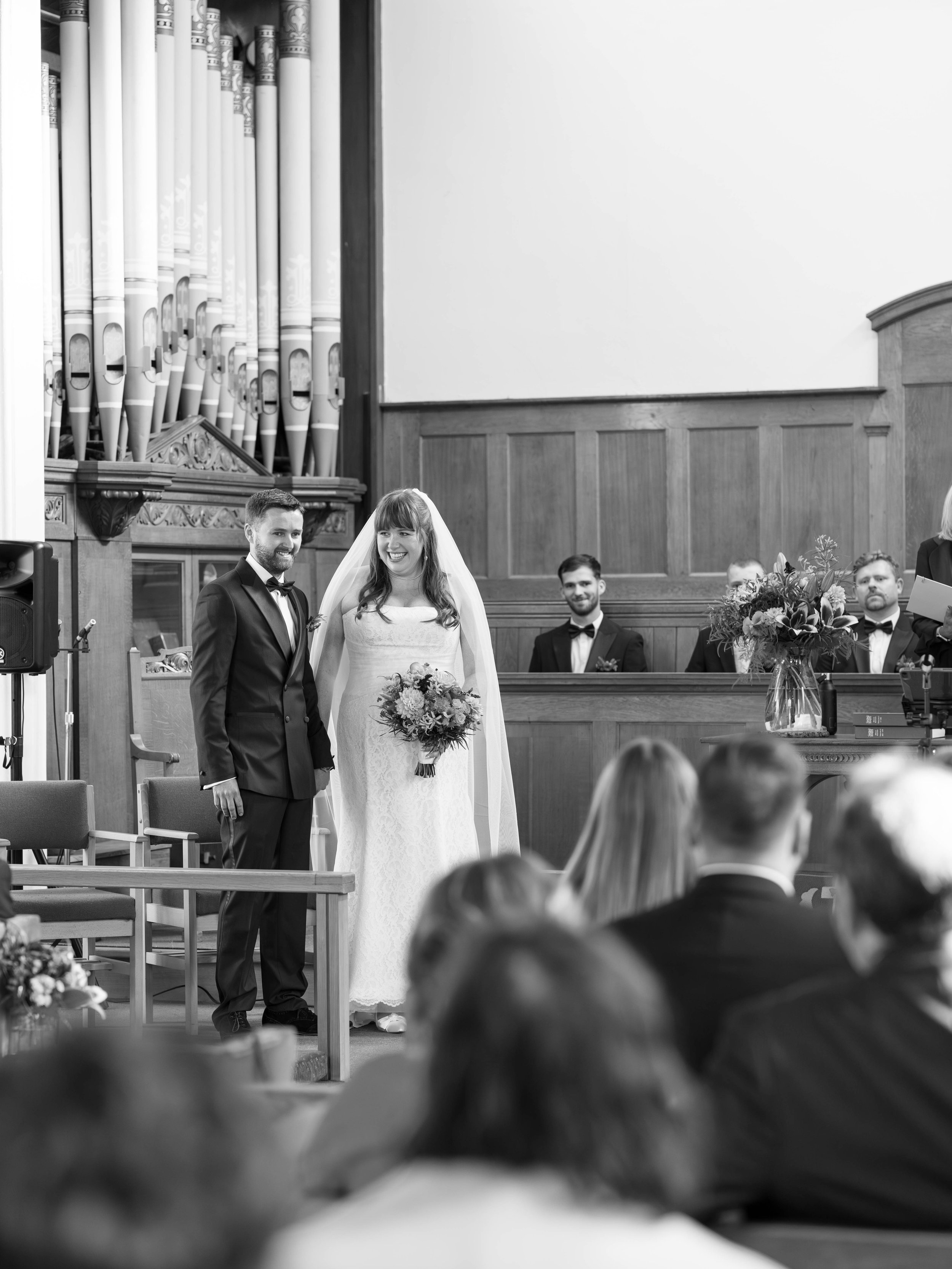 A black-and-white photo of a wedding ceremony with a bride and groom standing at the altar, surrounded by witnesses and possibly a minister, in a church with large organ pipes and wooden paneling.