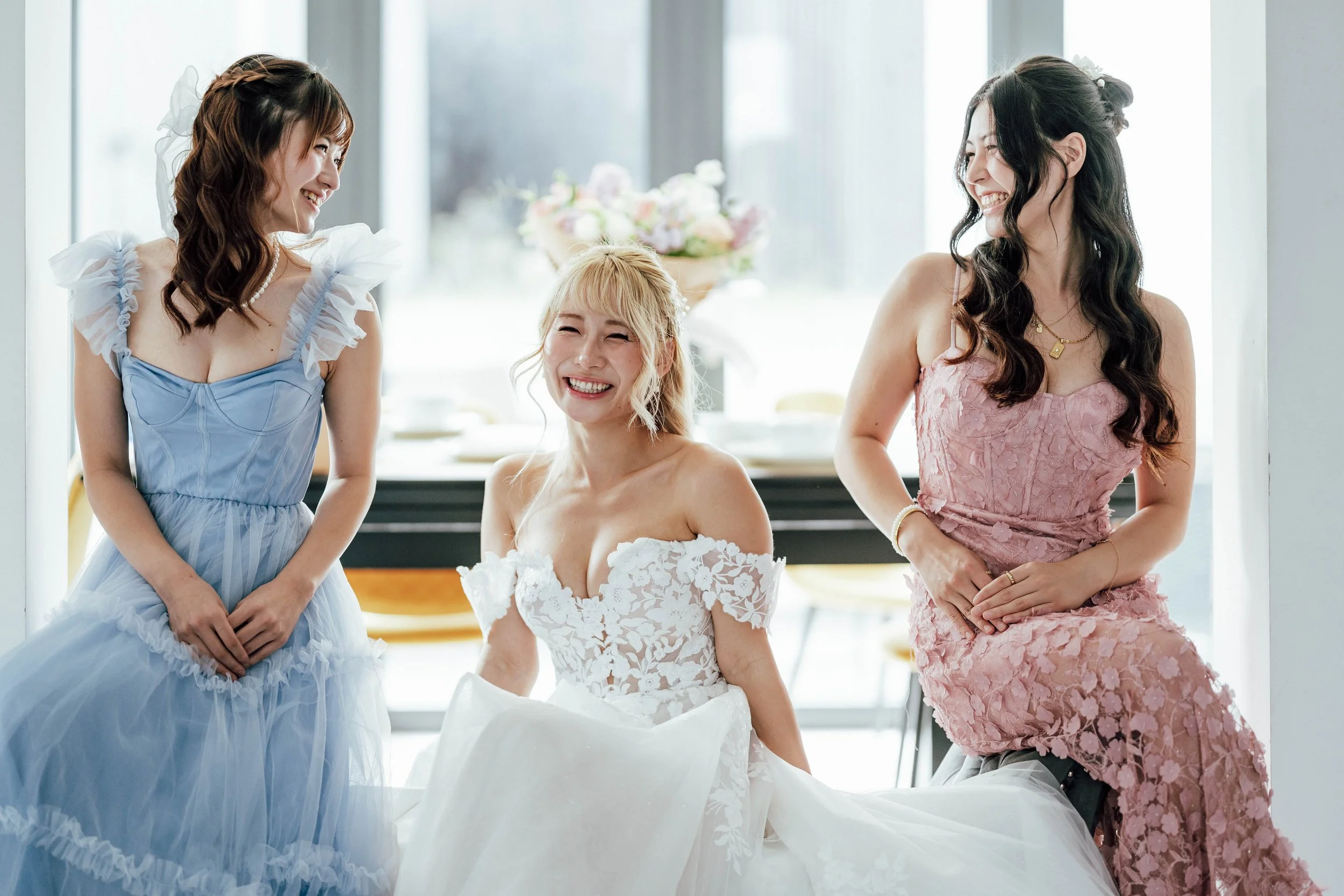 Three women at a wedding celebration, with the bride seated in the center wearing a white lace wedding dress, and two women in pastel-colored dresses standing and smiling beside her, in a bright room with large windows and a bouquet of flowers in the
