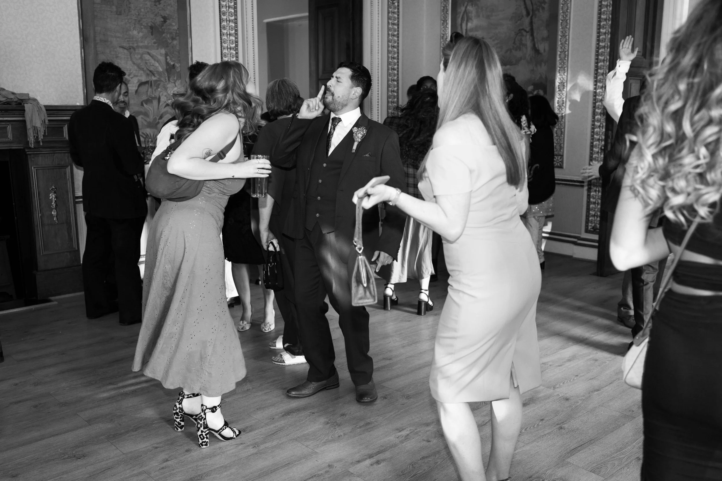 People dancing and socializing at a formal event or party in an elegant room with patterned walls and wooden flooring.