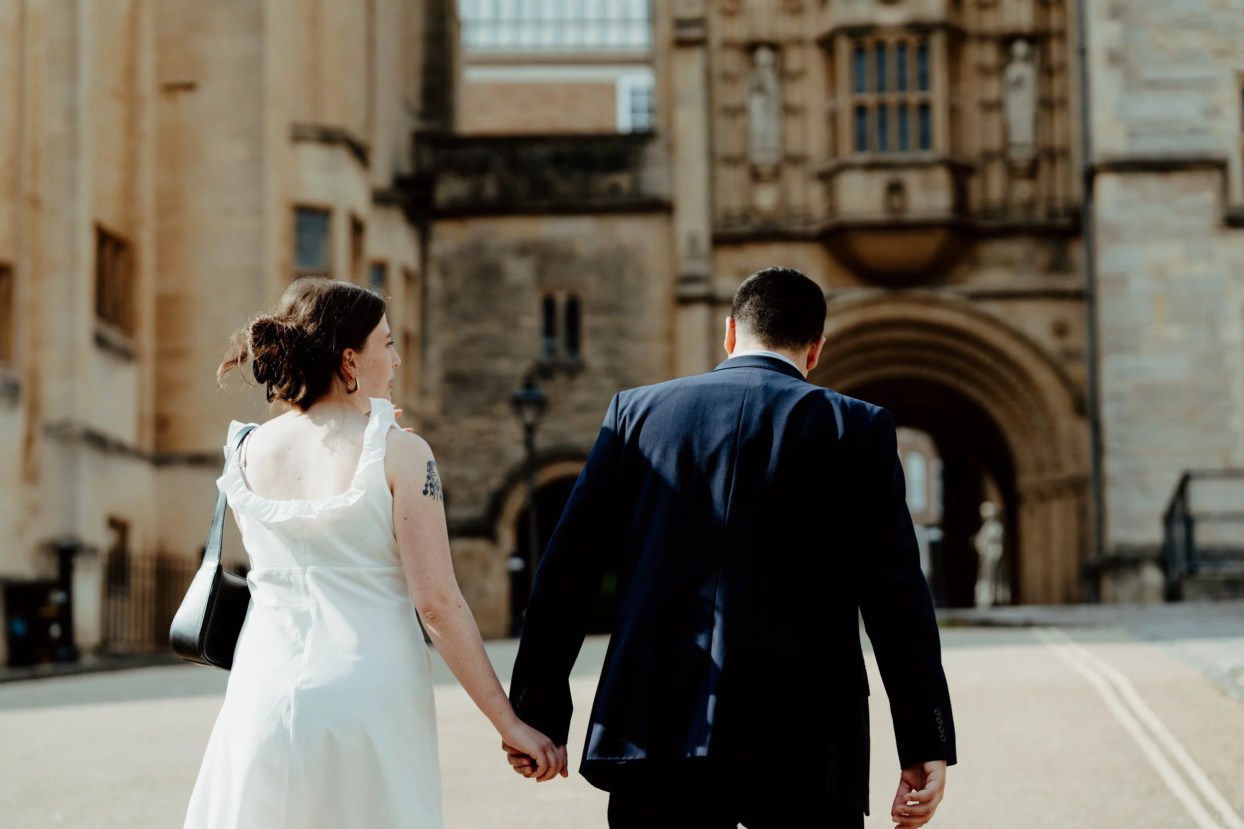 A woman and a man in formal attire walking hand in hand through a historical stone archway.