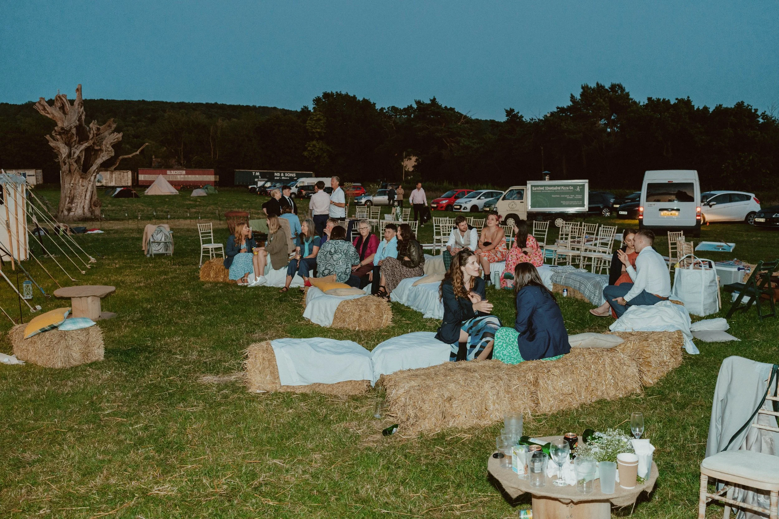People gathered outdoors on hay bales and folding chairs, socializing during an evening event in a grassy field with tents, trees, and parked cars in the background.