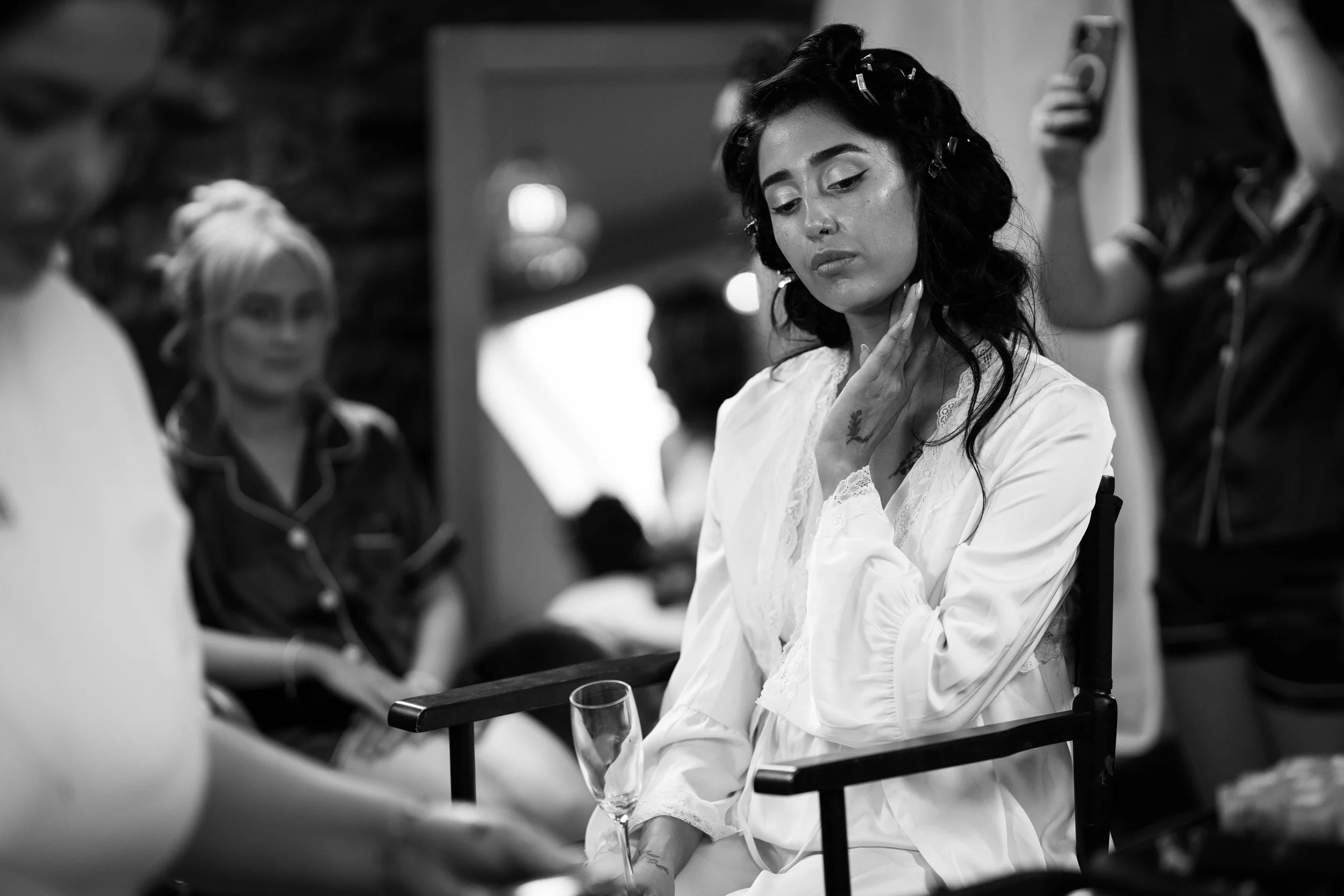 Black and white photo of a woman sitting with her hand on her neck, wearing a satin robe, with a glass of champagne on her lap, in a room with blurred people in the background.